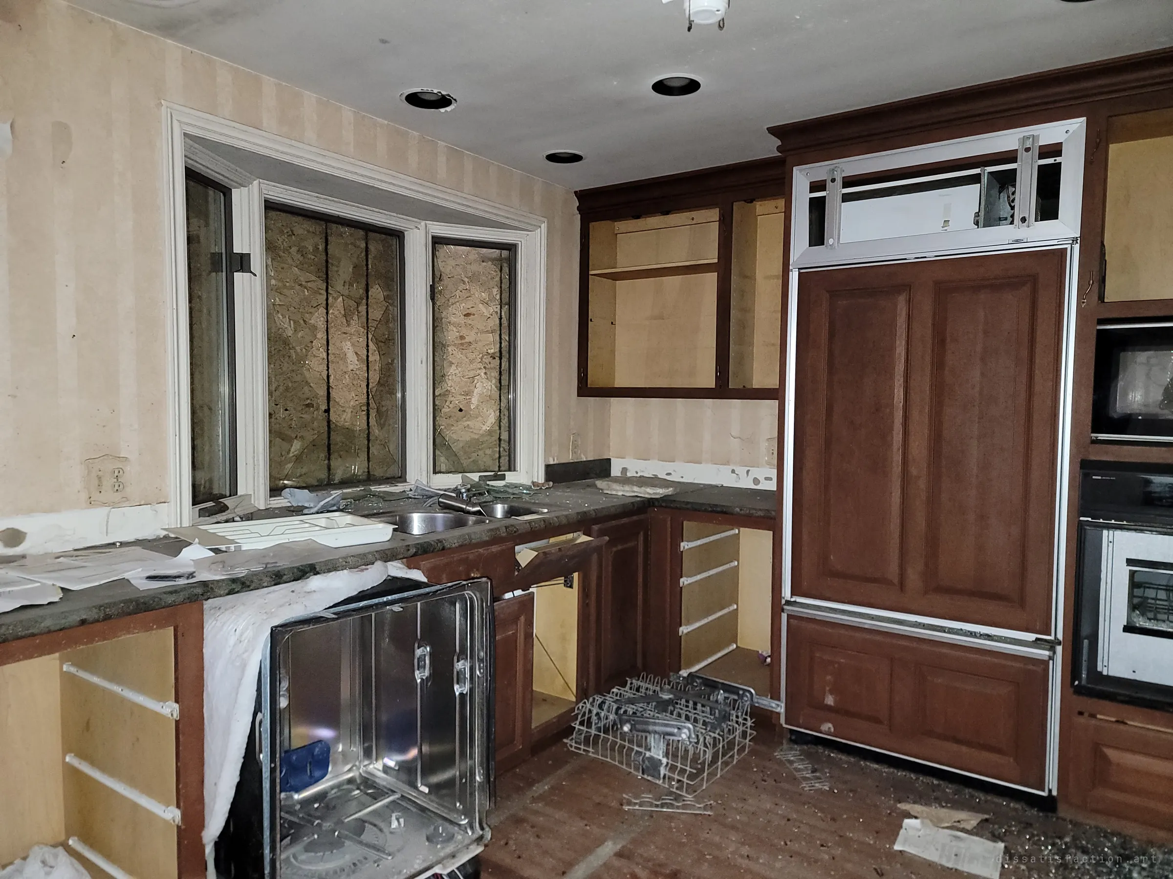 The back of the kitchen with an open dishwasher, its rack thrown on the floor, gutted cabinets and a boarded bay window above the sink. Scattered papers and glass show the room was ransacked.