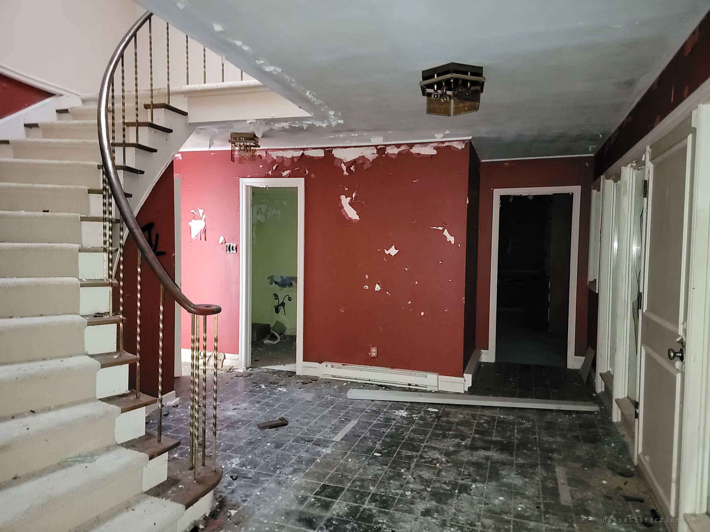 A front hall with a curving staircase and twisted brass balusters, red walls peeling in patches and debris across the dark tile floor. The grand entrance shows what the house once was.