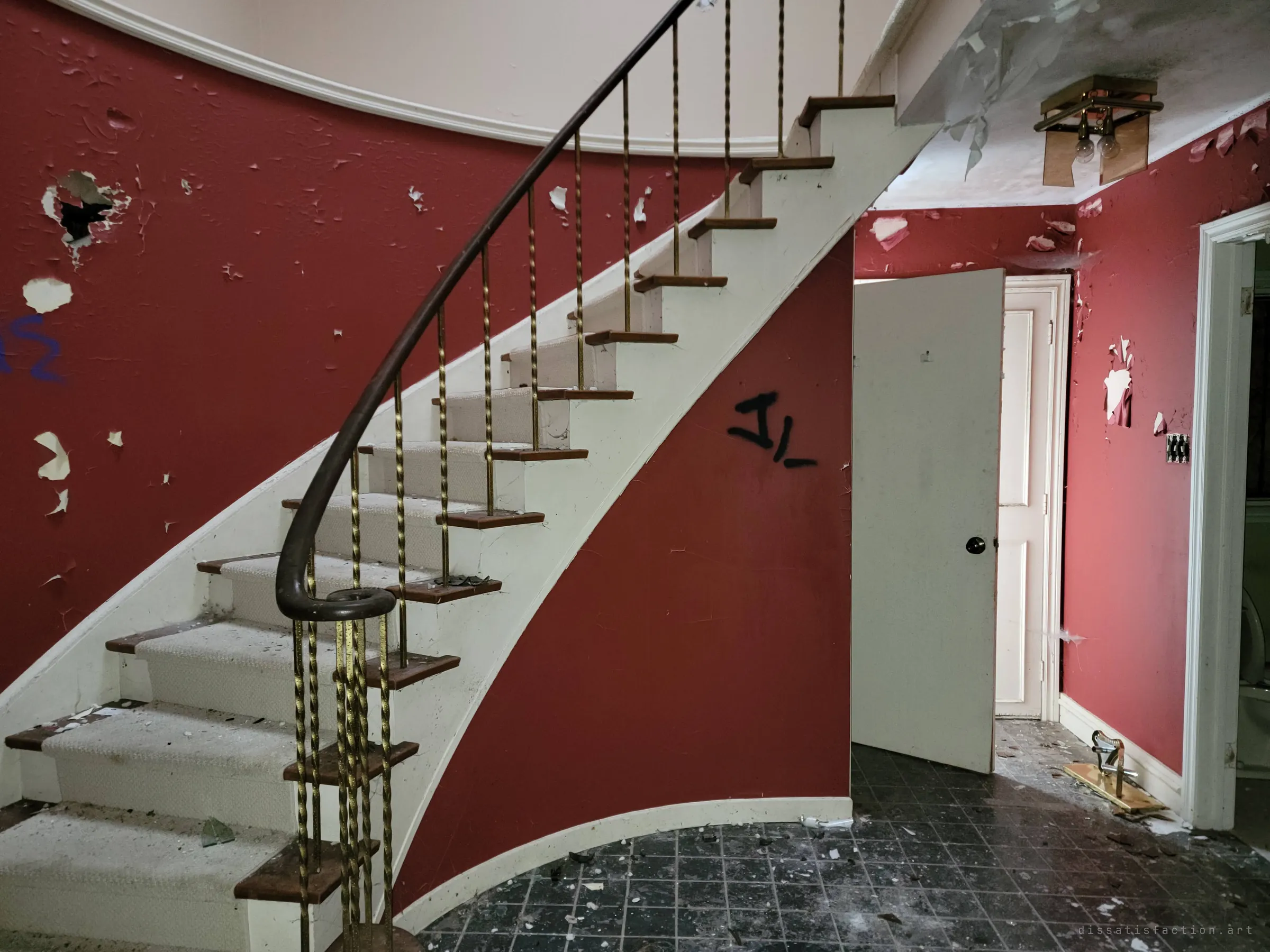 The curved staircase sweeping up along a red wall scarred with holes, peeling paint and a small spray-painted tag. Its elegant lines stand out against the damage around it.
