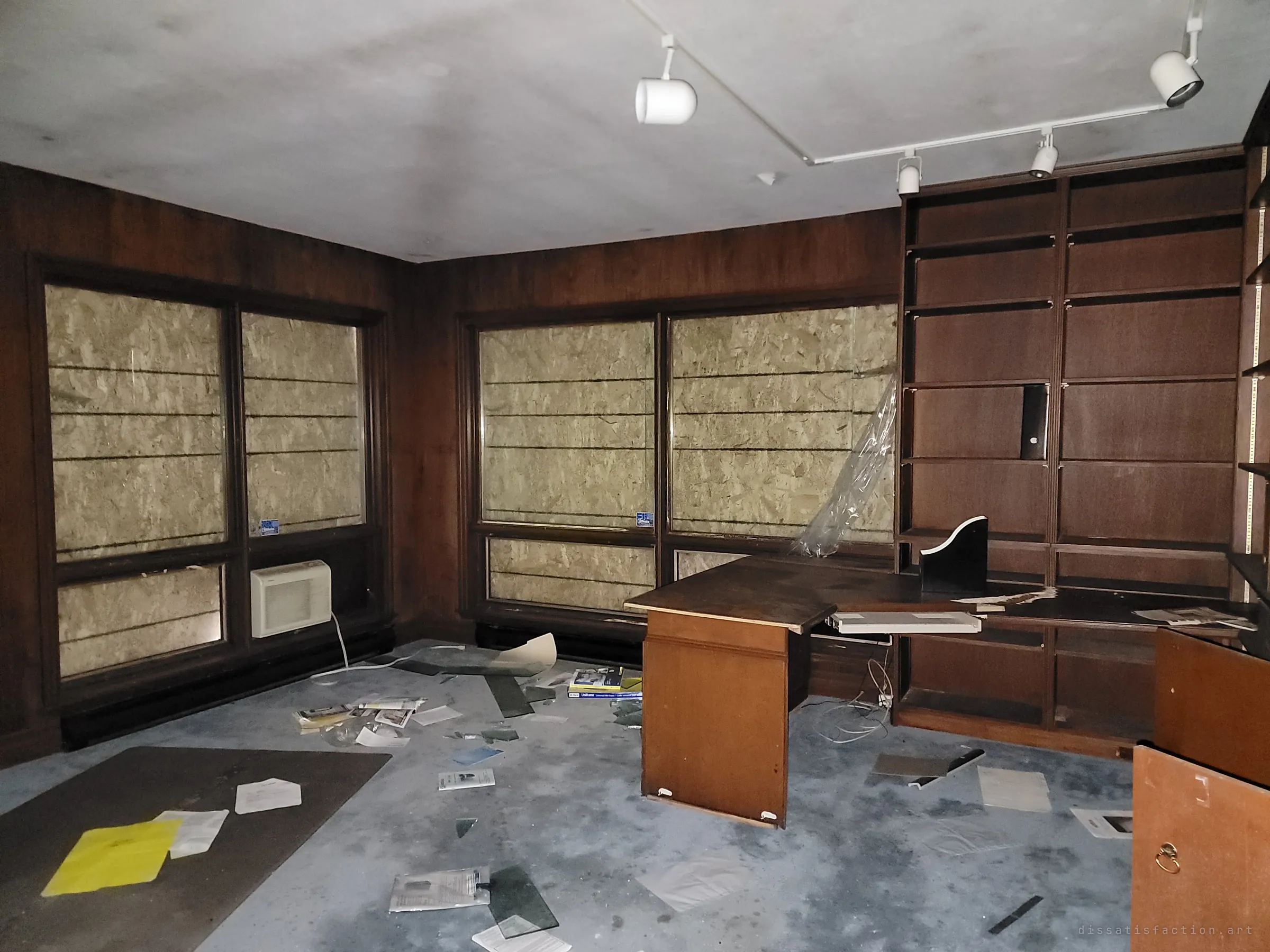 A wood-panelled study with boarded-up windows, empty built-in shelving and a desk pulled apart, the carpet strewn with broken glass and scattered manuals. The track lighting still hangs over a room that has been gutted by intruders.