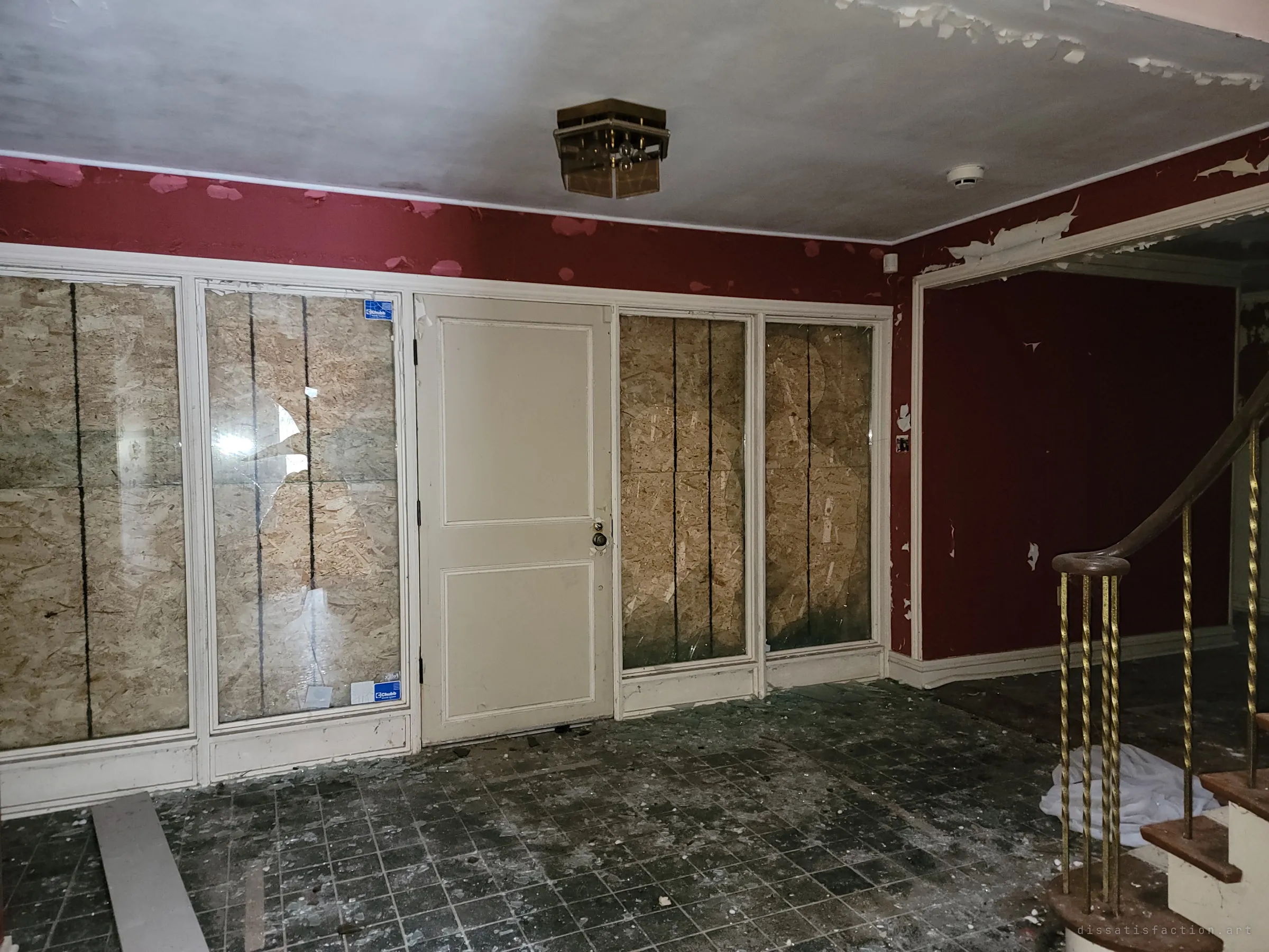 The front foyer: a panelled door flanked by smashed glass panes backed with plywood, red walls shedding paint and a twisted brass stair railing. Shattered glass covers the dark tile floor.