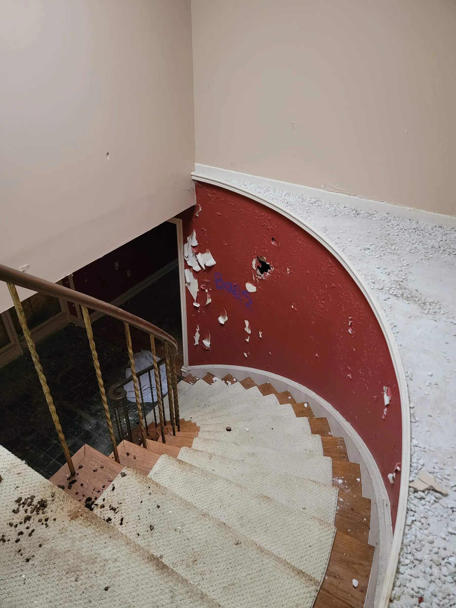 Looking down a curved carpeted staircase past a peeling red wall with a fist-sized hole and a scrawl of graffiti, toward the debris-covered foyer below. Gravel and dirt have been tracked across the landing.