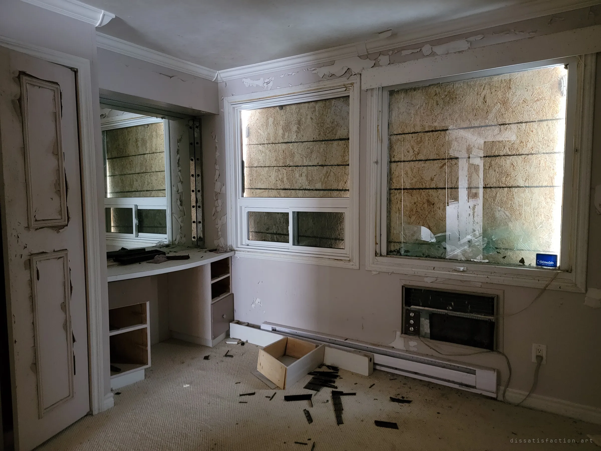 A dressing room with a built-in vanity, its drawers pulled and dumped, beside windows boarded from outside with light leaking around the edges. Paint curls off the doors and trim.
