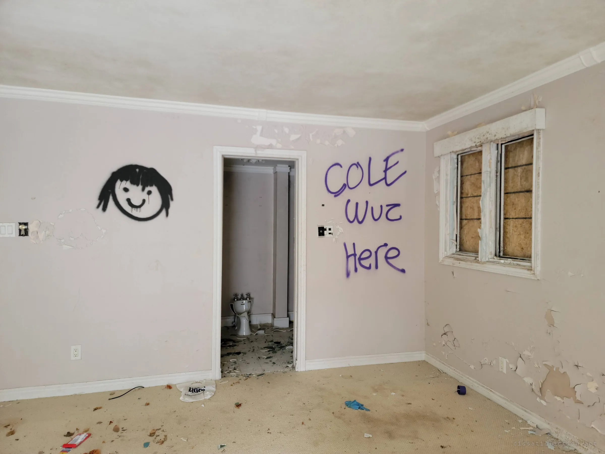 A pale bedroom tagged with a dripping cartoon face and a vandal's 'was here' message, the doorway opening onto a wrecked ensuite. The boarded window and peeling walls show how far the house has fallen.