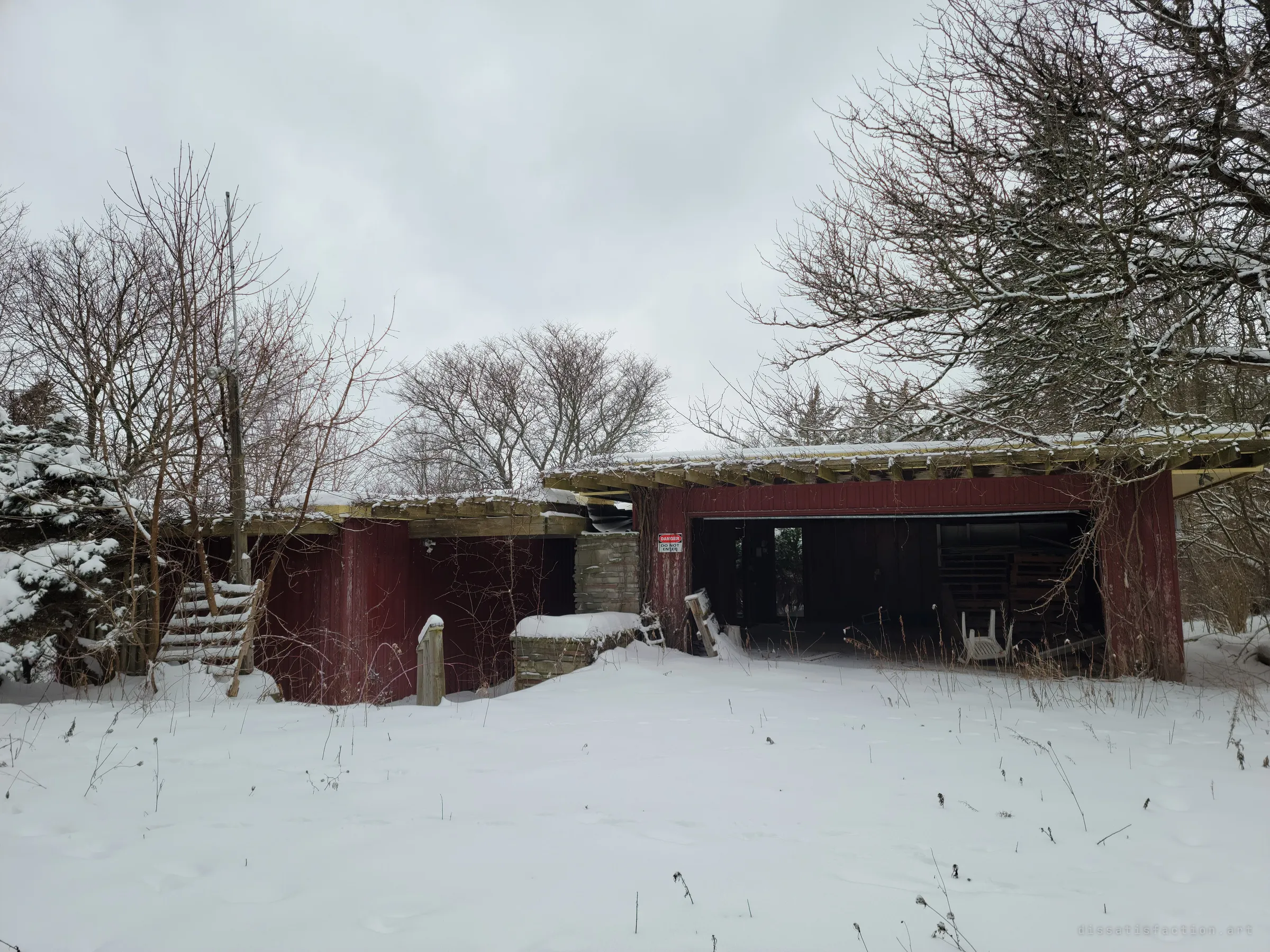 A snowed-in, low red house with open timber rafters and a gaping garage, a danger sign by the entrance. The first look at a mid-century home left to the weather.