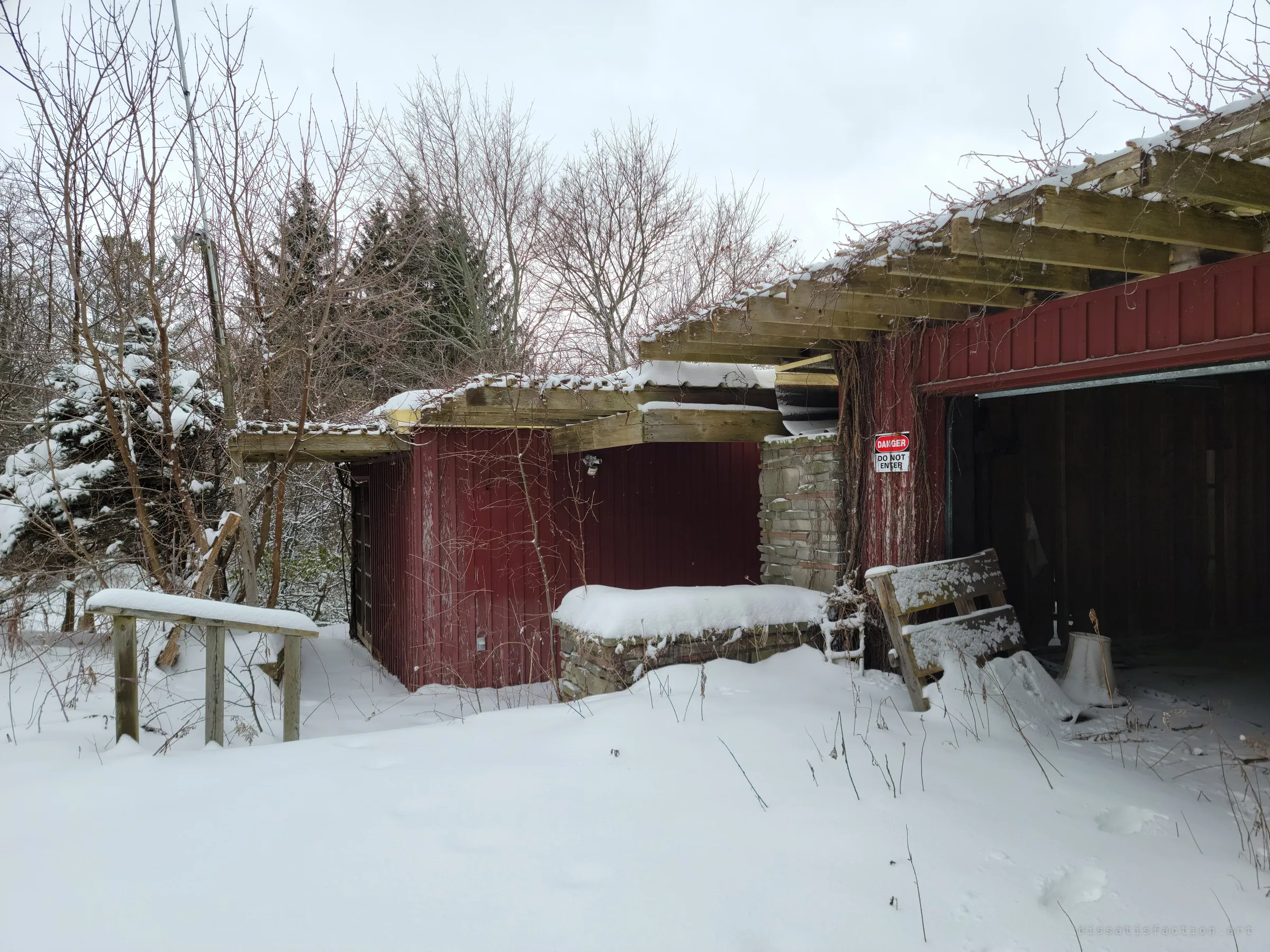Snow drifts against the red siding and stone planter at the front of the house, rotting rafters overhead and a generic danger sign beside the open garage.