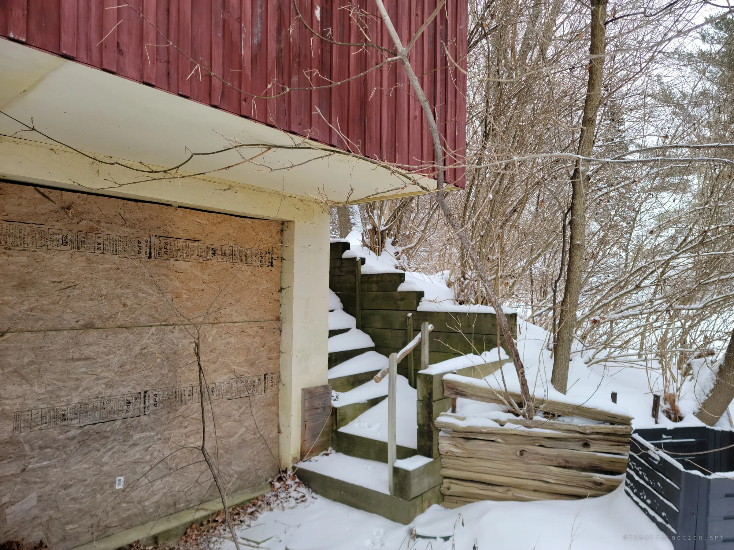 A garage door boarded with plywood under the red upper storey, timber steps climbing a snowy slope beside it.