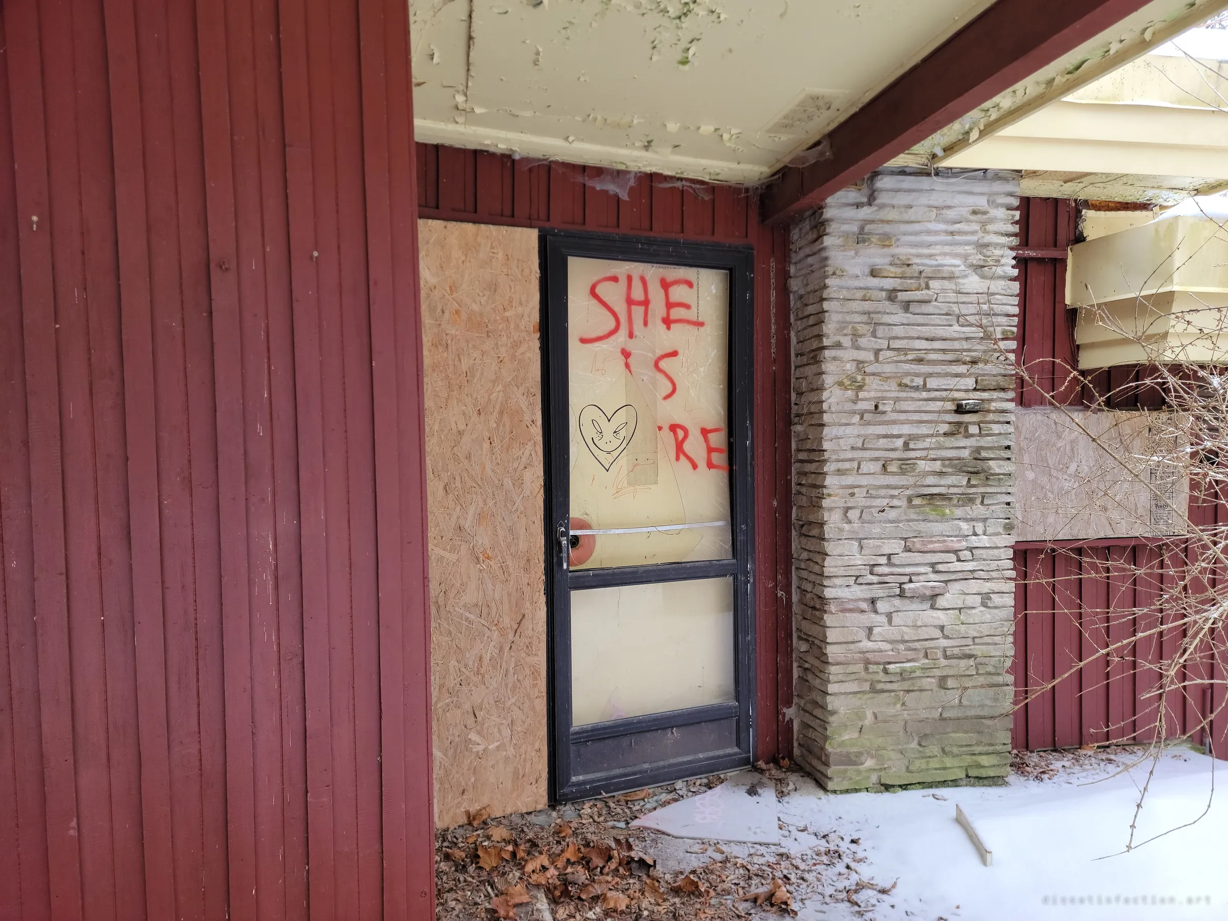 A glass storm door sprayed with red graffiti and a doodled heart face, flanked by plywood and a stacked-stone pillar under a peeling porch ceiling.