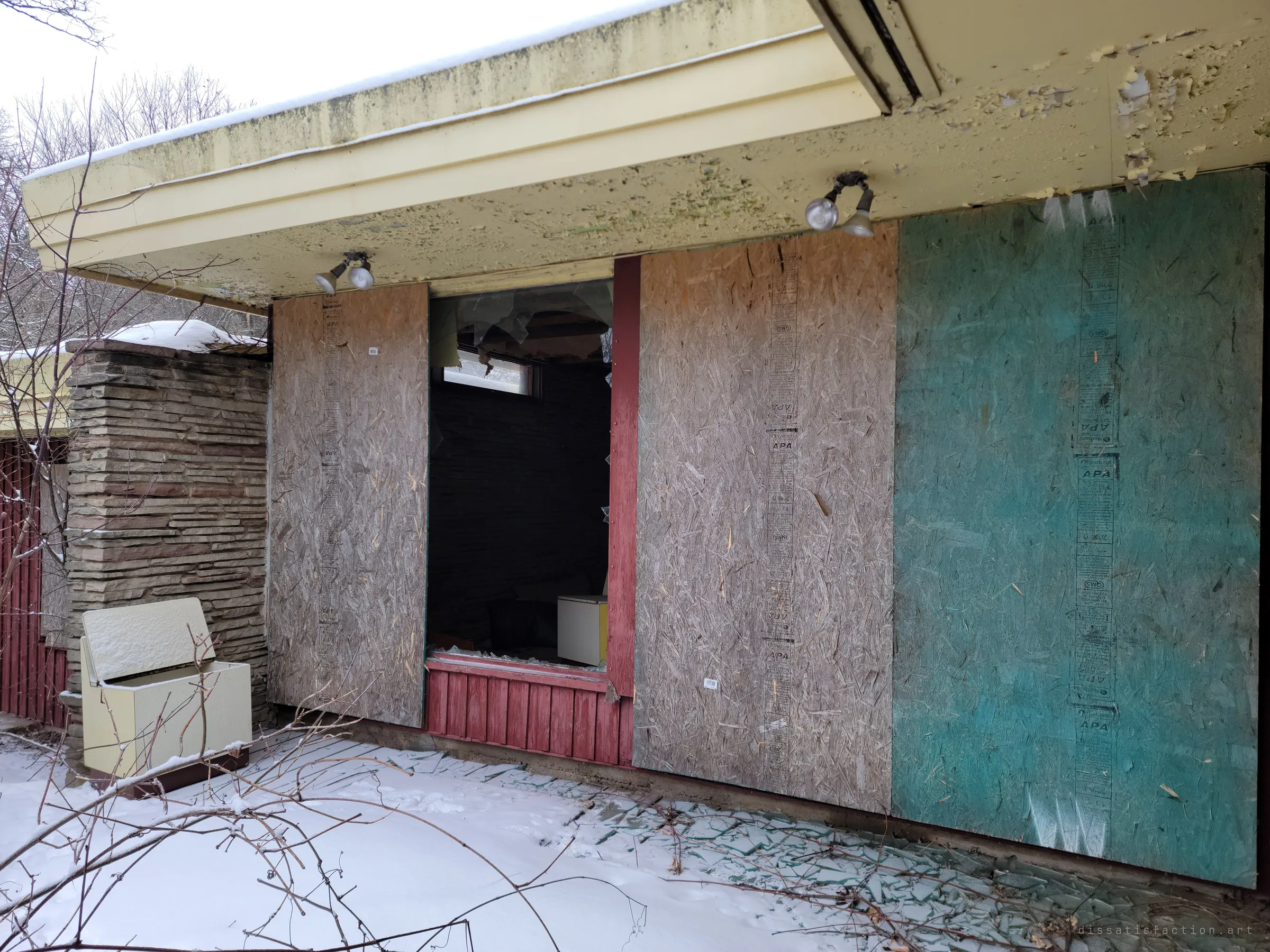 Boarded windows along the back wall, one pane smashed open onto a dark room, shattered glass scattered across the snow below.