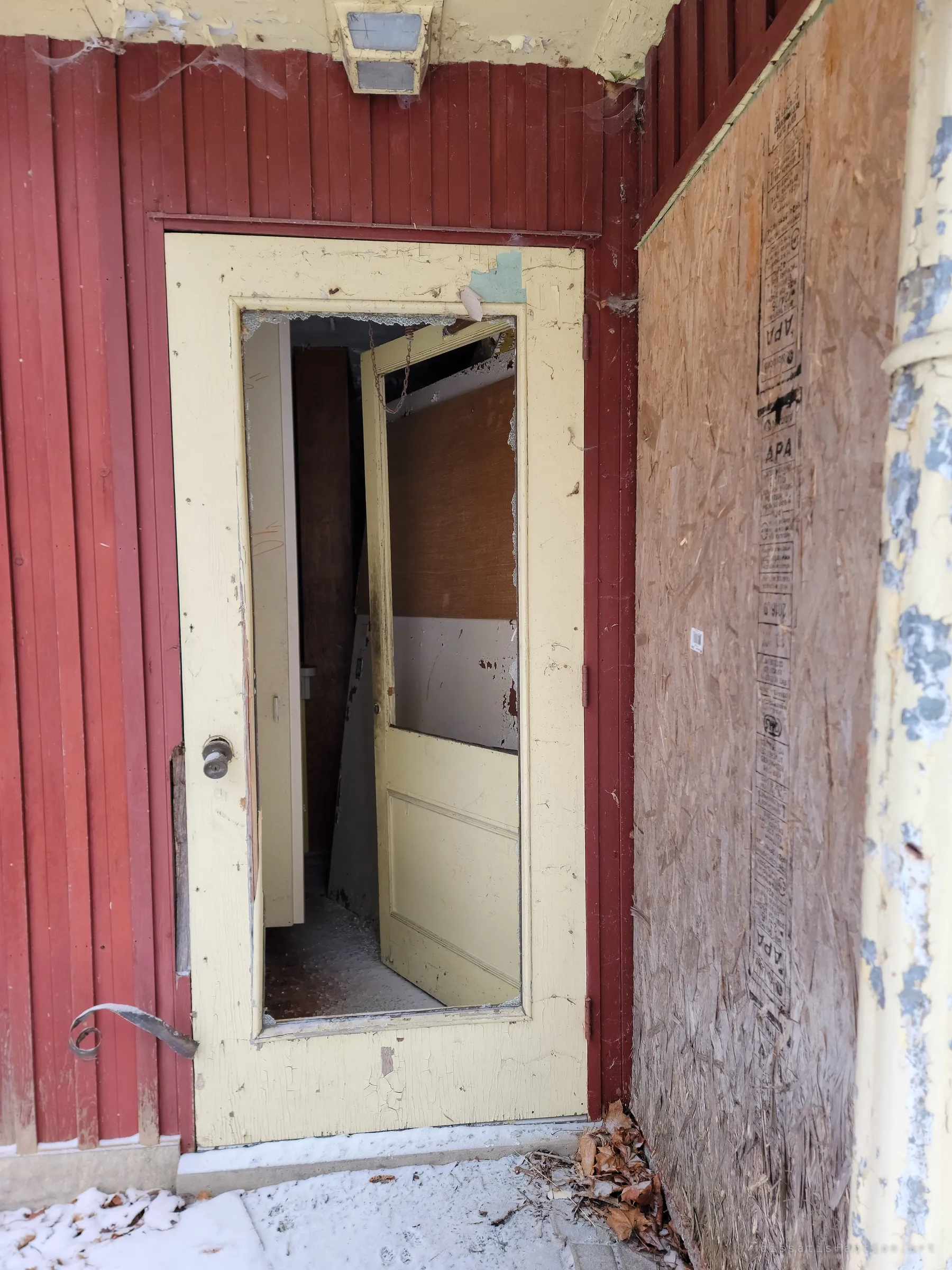 A cream door with its glass knocked out, showing another door hanging open inside, set in red siding next to a boarded window.