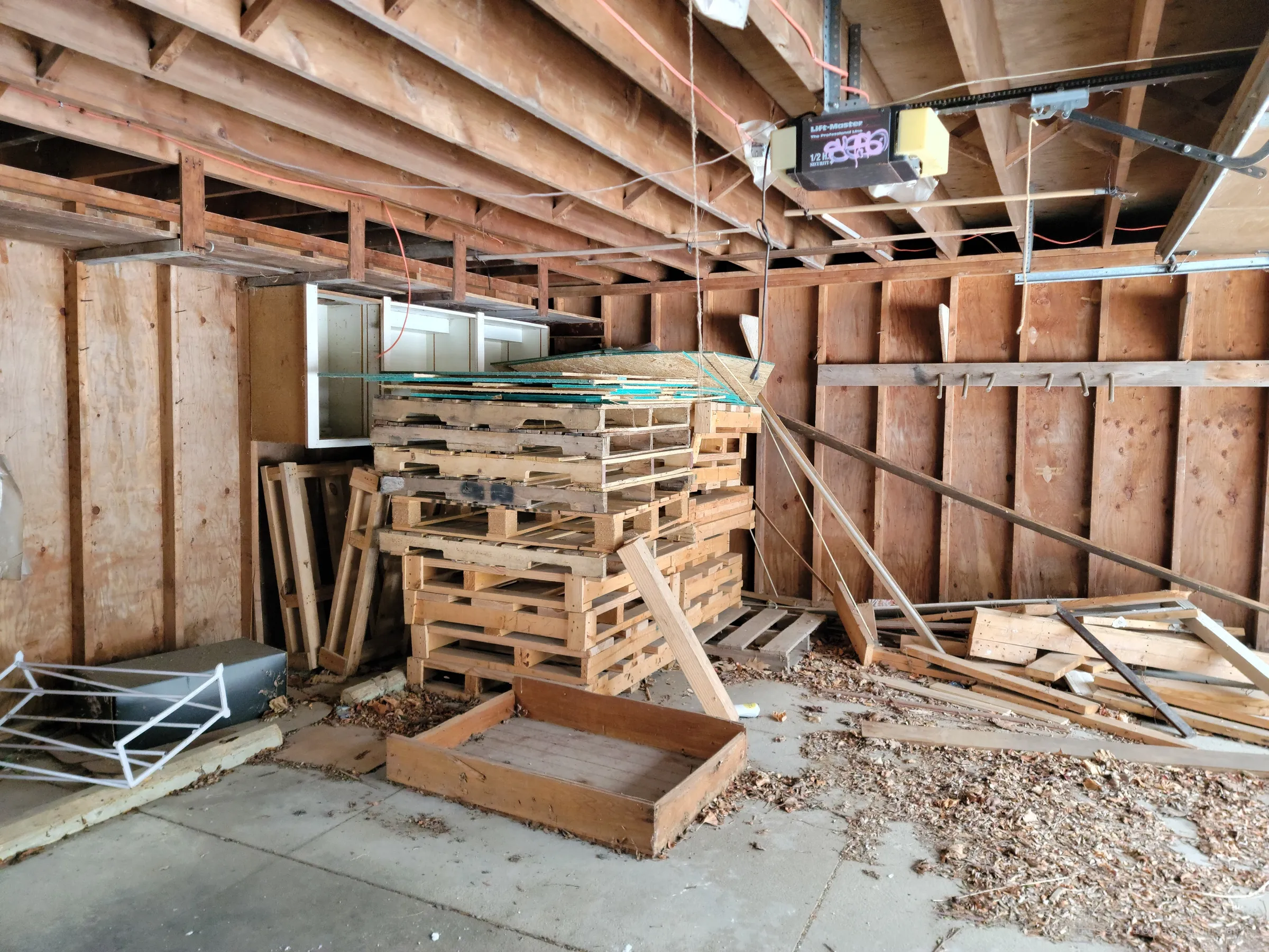 The stripped garage interior: stacked pallets, loose lumber and a tagged door opener hanging from bare joists.