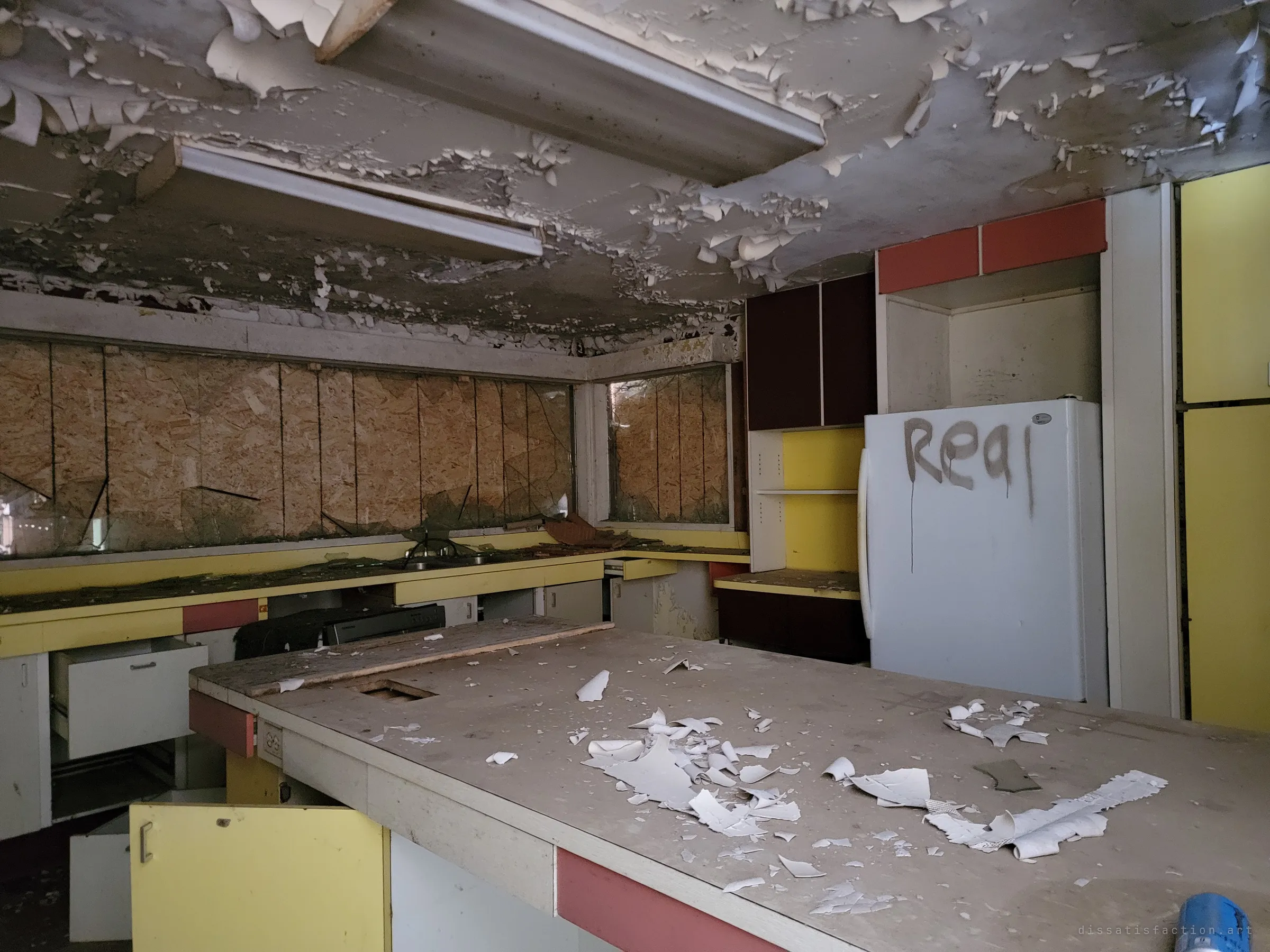 A retro kitchen in yellow and coral laminate under a ceiling of curling paint, the island strewn with paint flakes and the windows boarded behind shattered glass. A graffitied fridge stands in the corner.