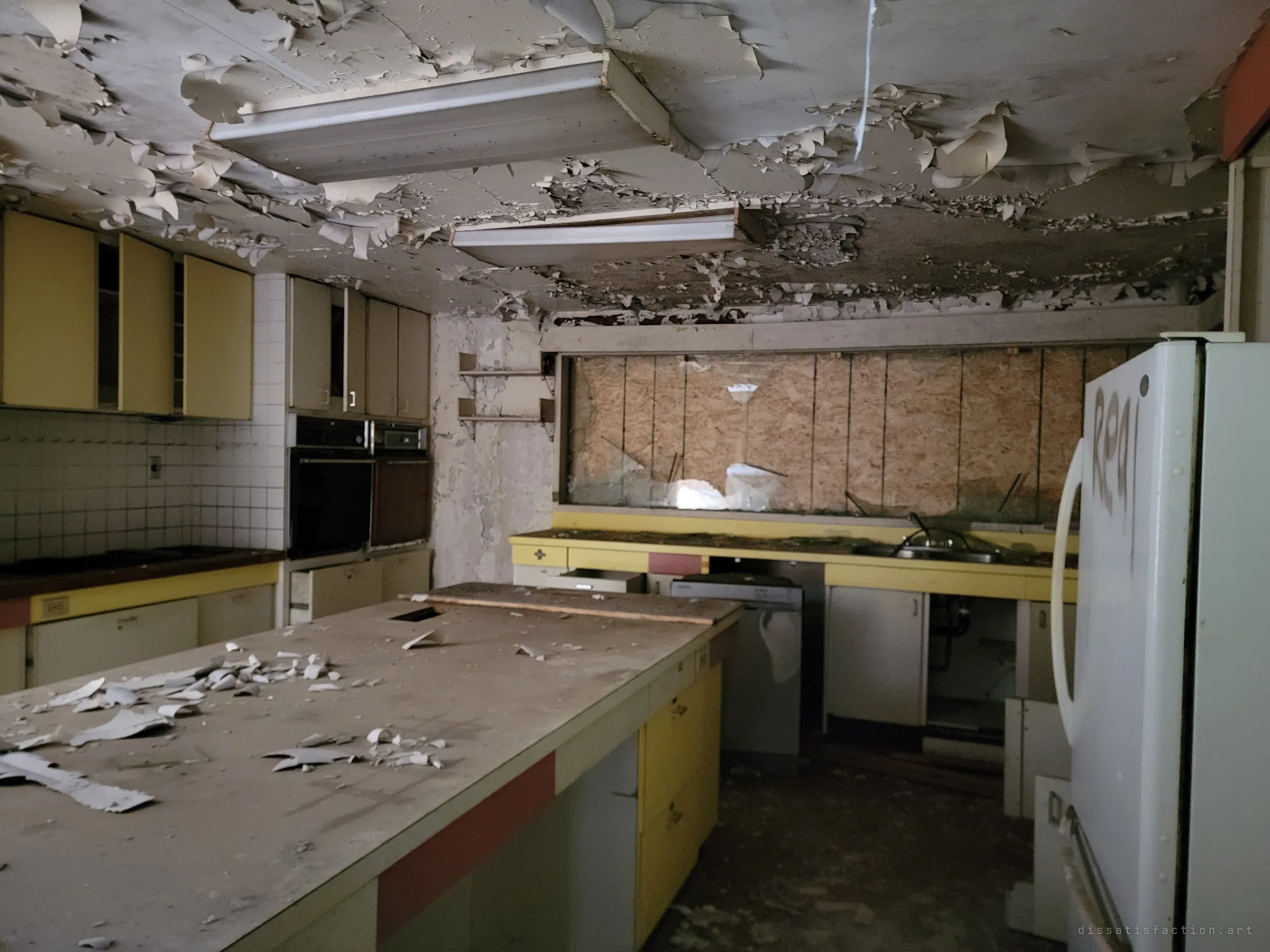 The same kitchen from the other end: built-in wall ovens, a tiled backsplash and a long island under peeling ceiling paint, with the tagged fridge looming in the foreground.