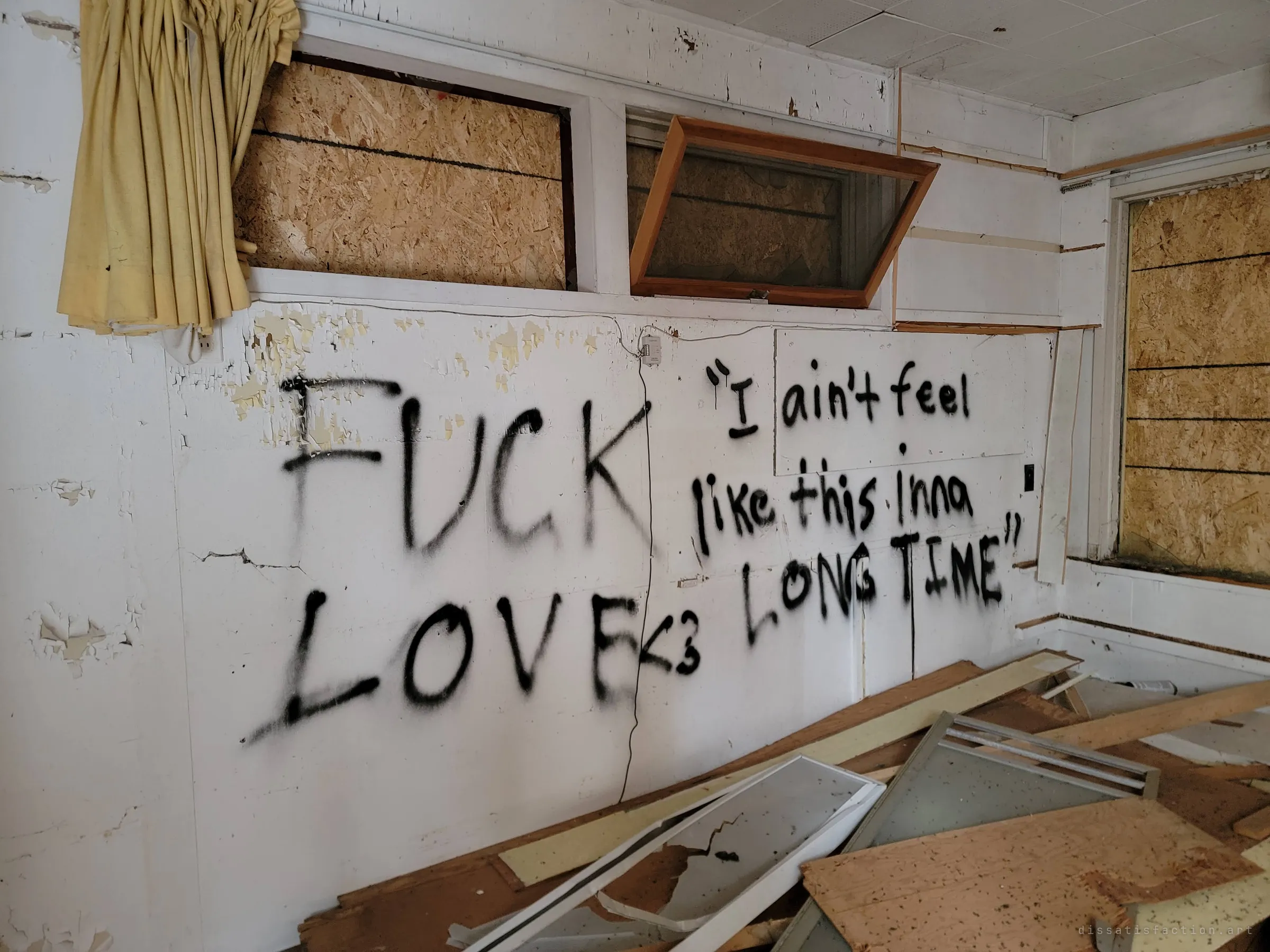 Black spray-painted lines about love and a long time cover a white wall under boarded windows and a faded yellow curtain, with broken mirror doors piled at its foot.