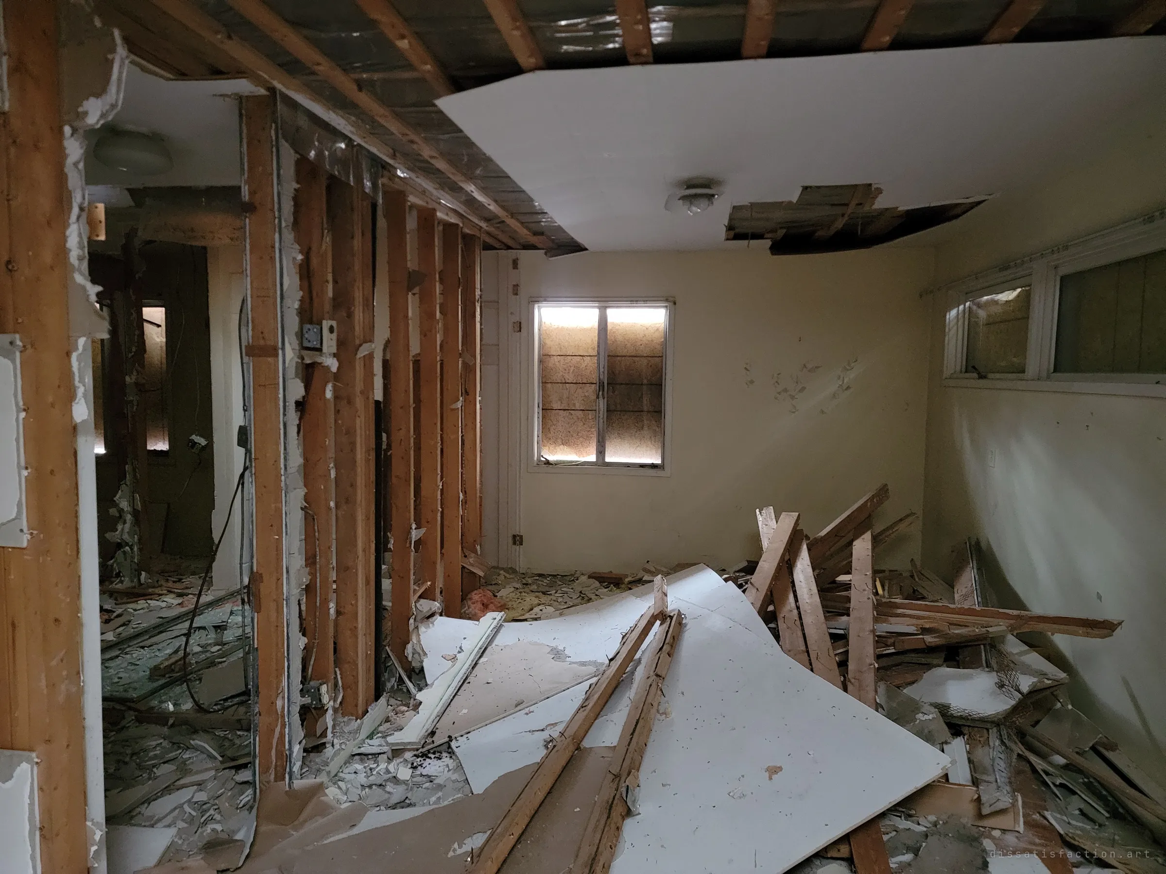 A bedroom torn open to its studs, with sheets of drywall and broken lumber heaped on the floor and daylight glowing around a boarded window.