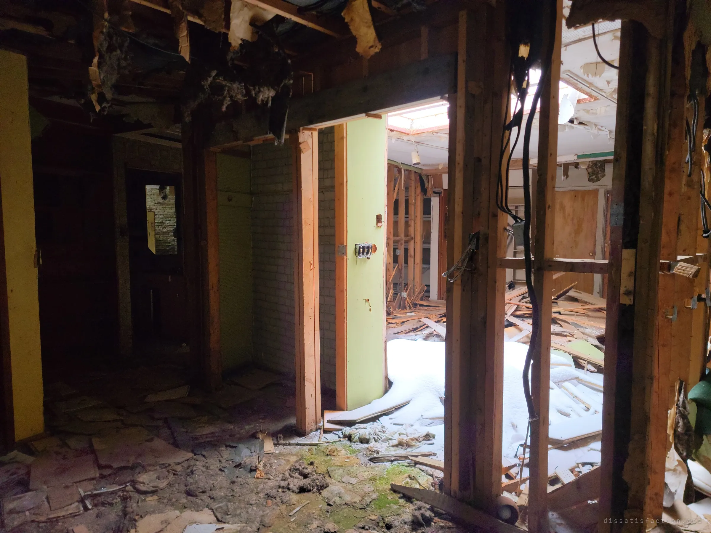 A dark hallway of stripped framing and dangling cables opens onto a bright room where snow has drifted in over the debris through the broken roof.