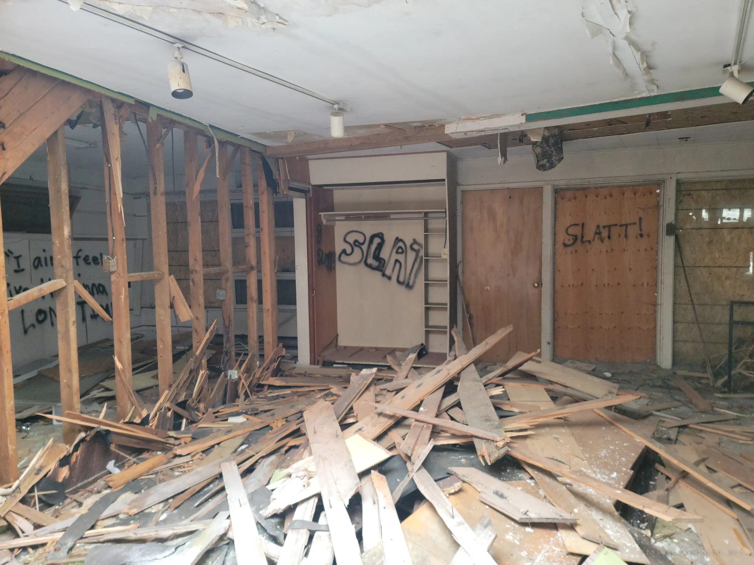 A room buried in splintered floorboards, its closet and doors tagged in black spray paint beneath a track of gallery-style spotlights.