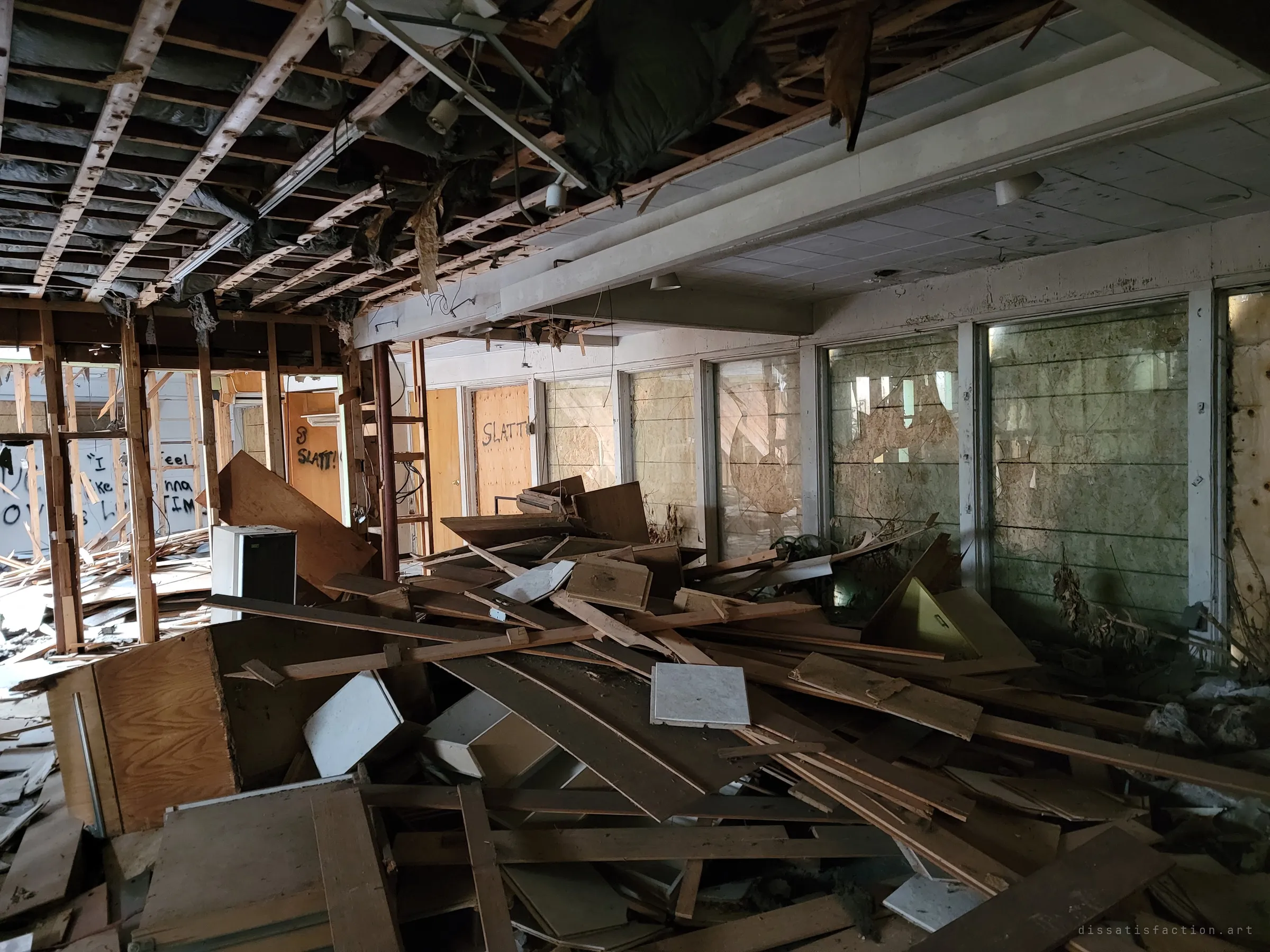 A long living space with its ceiling pulled down to the joists, a heap of cabinets and boards in the middle and a wall of boarded picture windows running along the right.