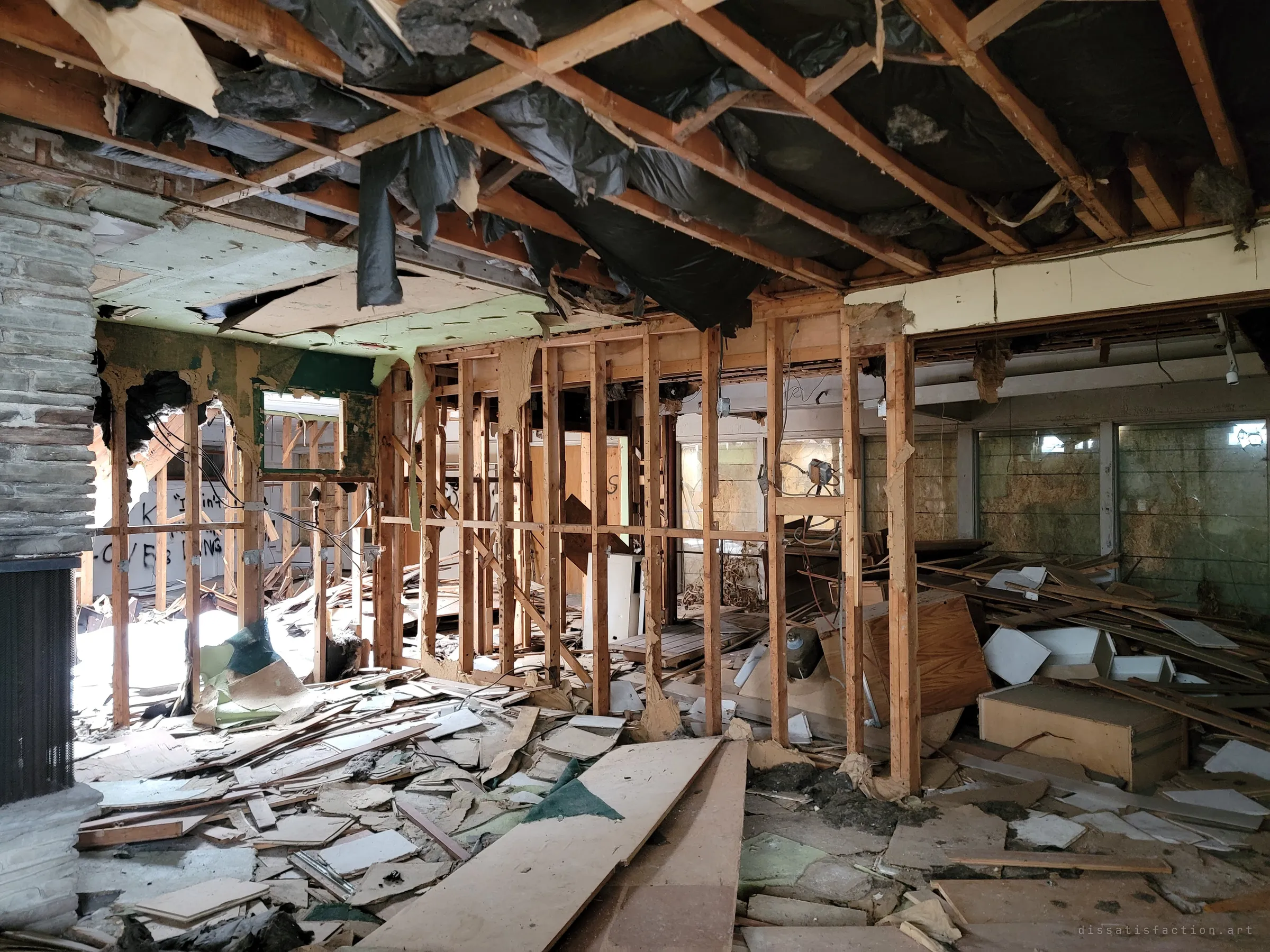 Beside a stacked-stone fireplace, the interior walls have been stripped to bare studs so the whole gutted floor reads through them, with insulation sheeting sagging from the open ceiling.