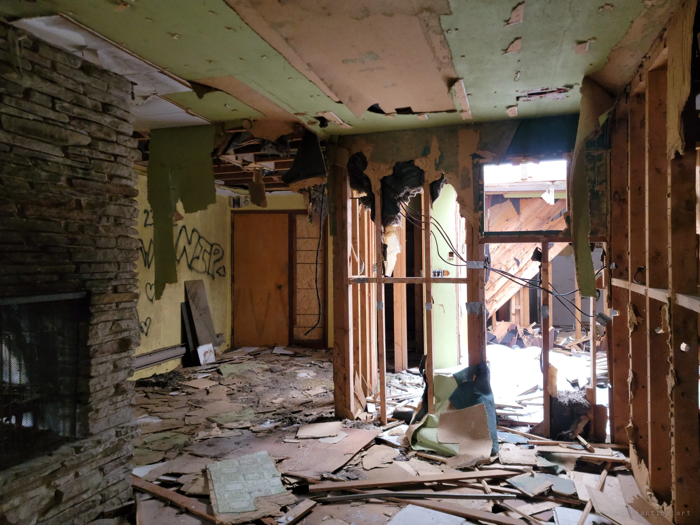 A stone fireplace and graffiti-marked yellow walls frame a gutted passage, where torn green ceiling board hangs in strips and daylight falls through a collapsed section beyond.