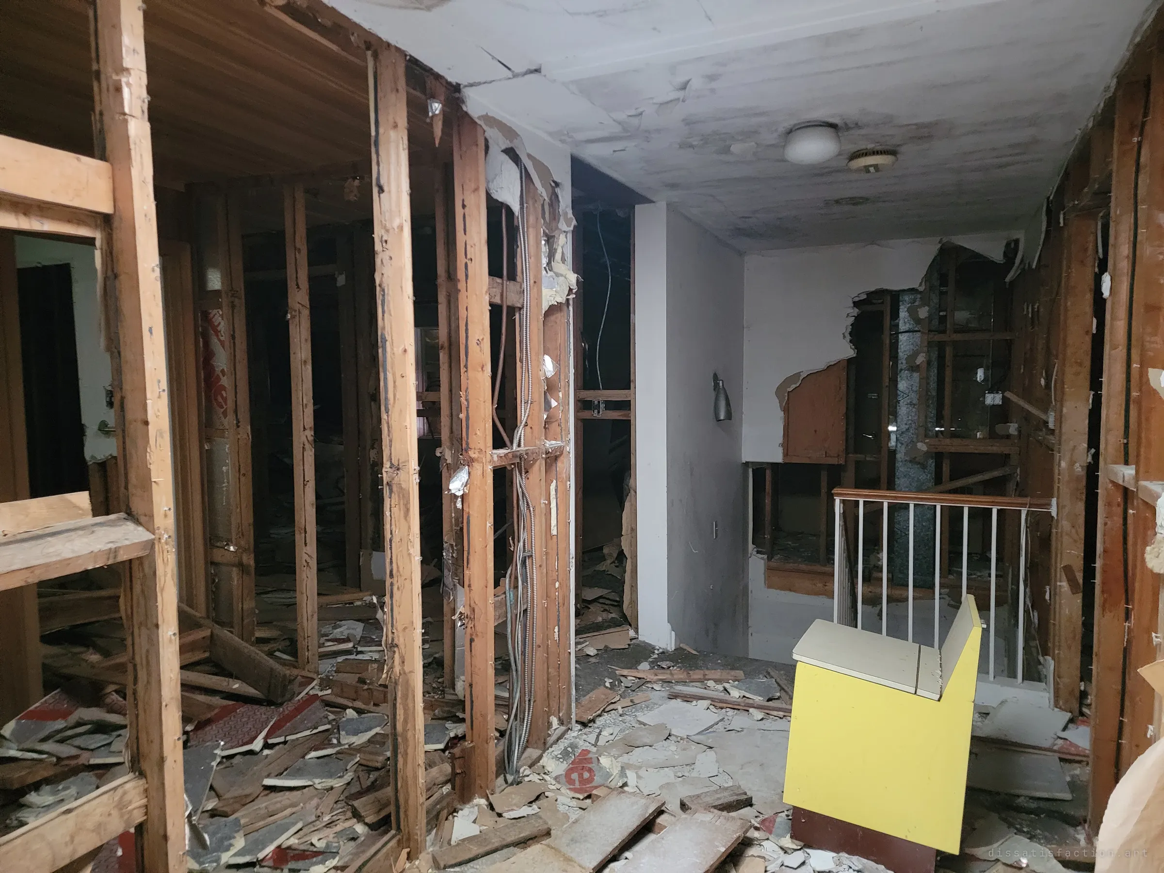 An upper hallway gutted to the studs, wiring hanging loose and flooring torn up around a lone yellow bench by the stair railing. Shows how far the strip-out had gone.