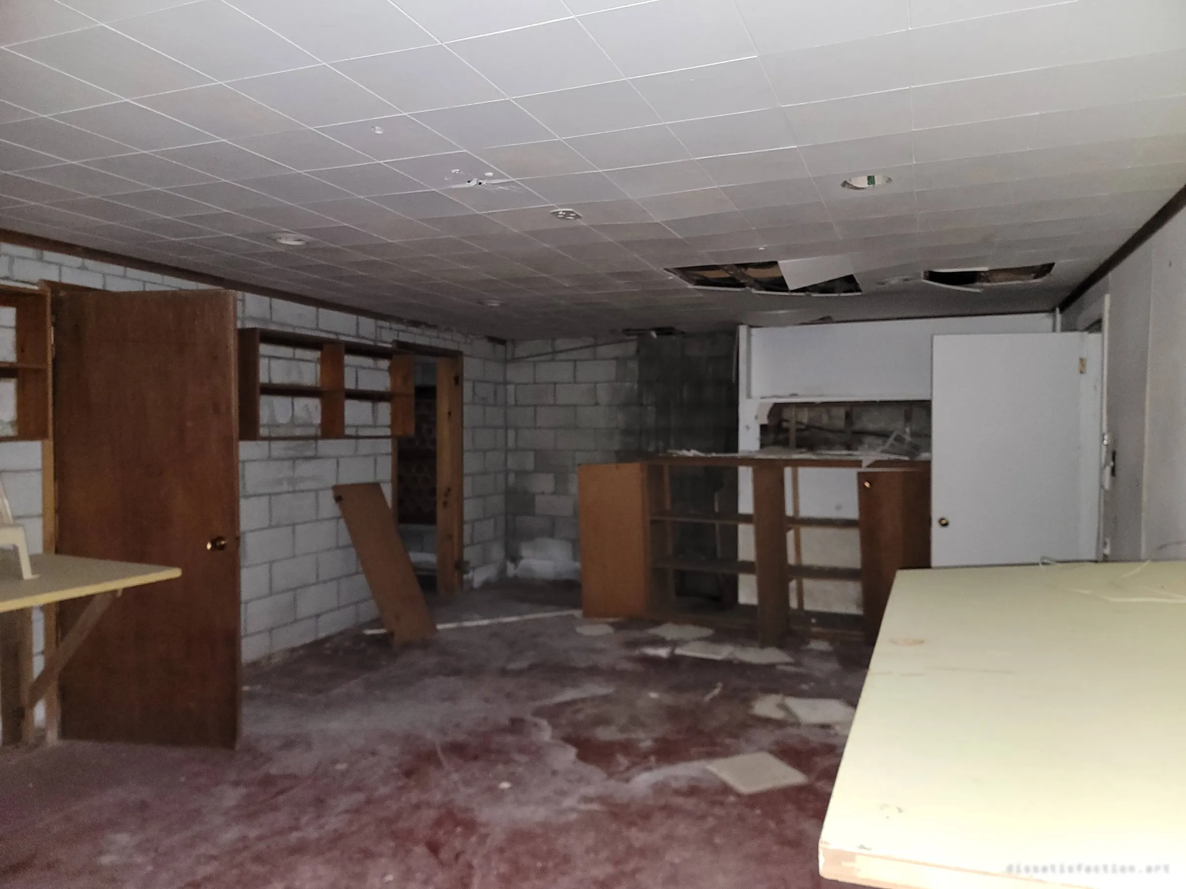 A basement rec room with a cracked drop ceiling, cinder-block walls and empty built-in shelving over a stained carpet. The leftover furniture hints at the family room it once was.