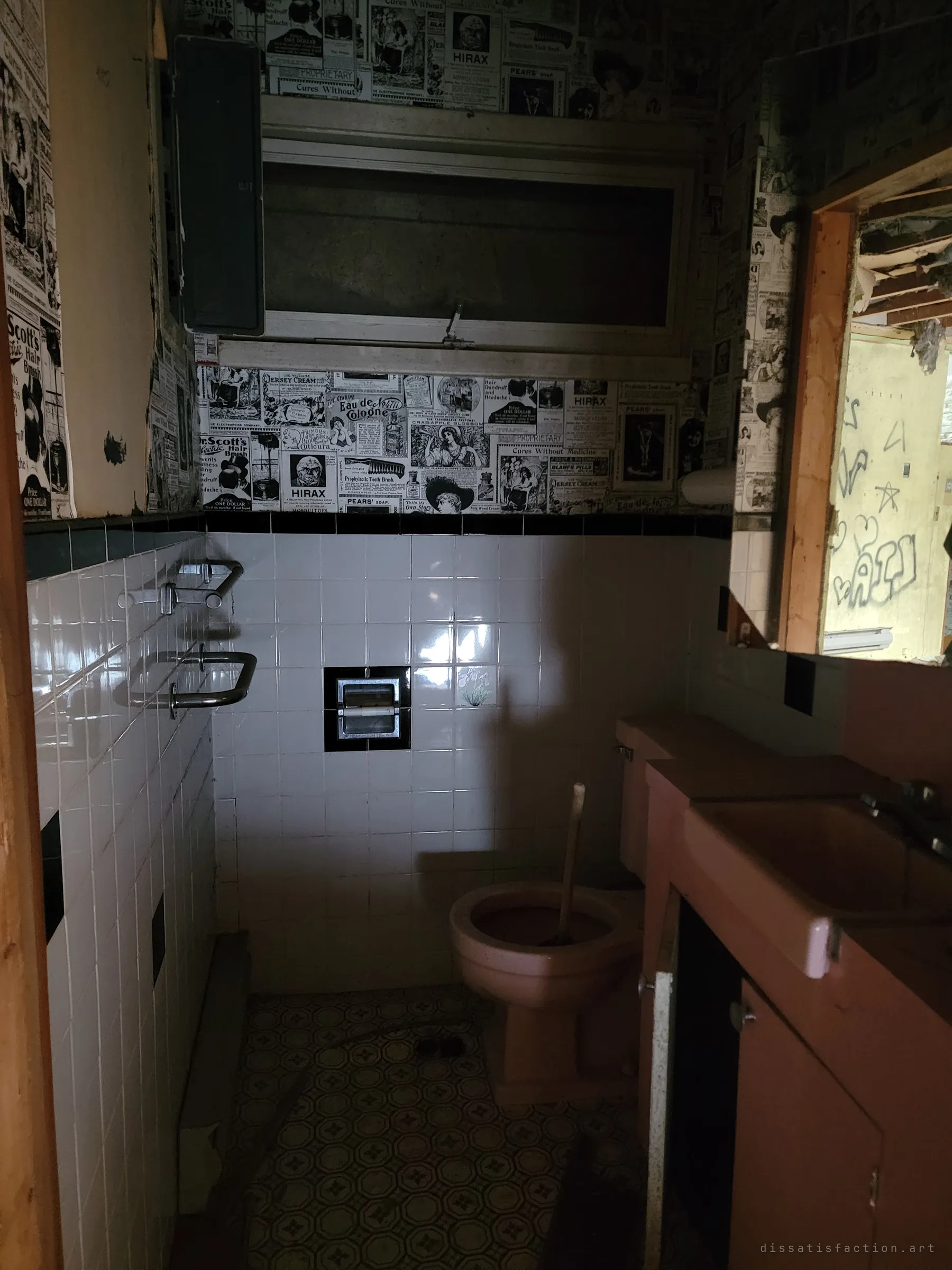 A small bathroom with pink fixtures under vintage newspaper-ad wallpaper, with graffiti visible through the doorway in the mirror. The retro decor sits against the later vandalism.