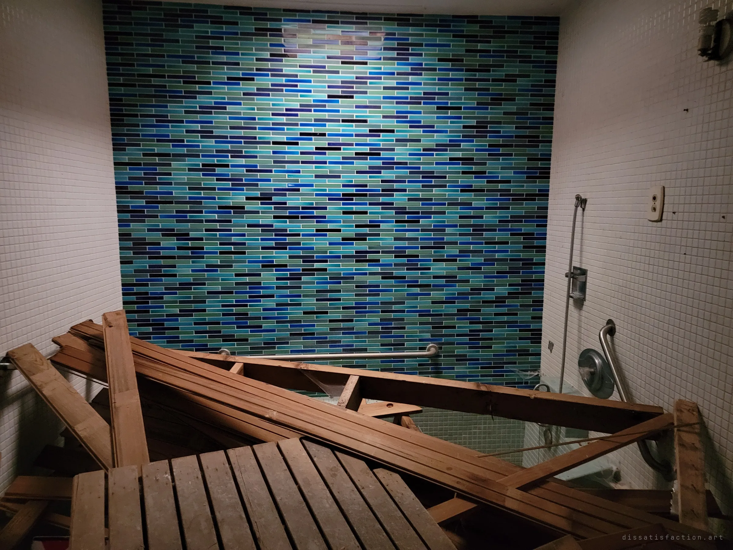 A sunken tub room with a shimmering blue glass-tile wall, now filled with a heap of discarded lumber. A striking finish half buried in debris.