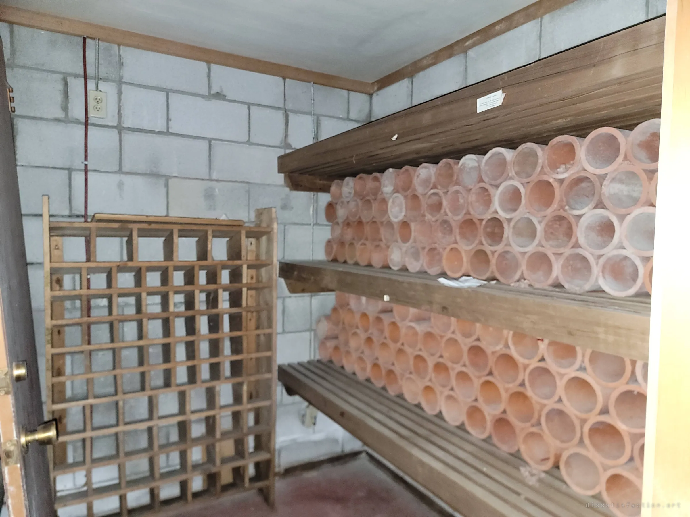 A cinder-block storage room with shelves stacked full of terracotta tubes beside an empty wooden cubby rack. An odd stash left behind in the basement.
