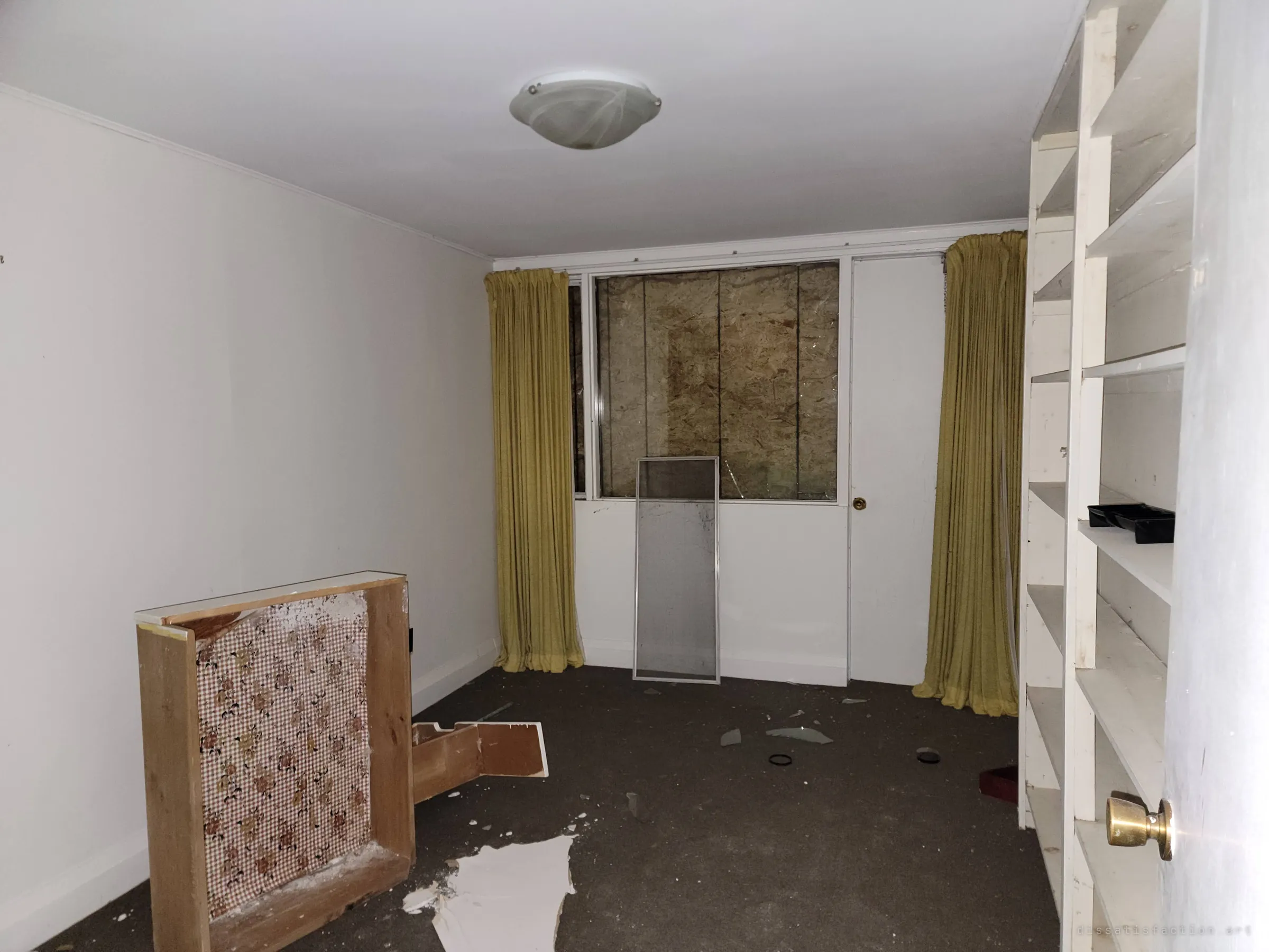 A bare bedroom with mustard curtains framing a boarded-up window, a broken drawer and glass on the carpet. The boarding shows the house being sealed up.
