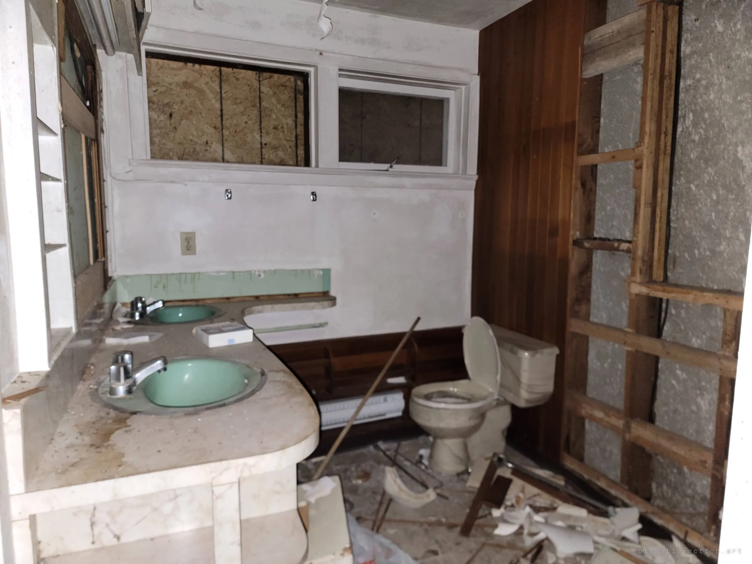 A wrecked bathroom with green double sinks, a boarded window and smashed ceramic across the floor. The torn-out wall exposes the framing behind.