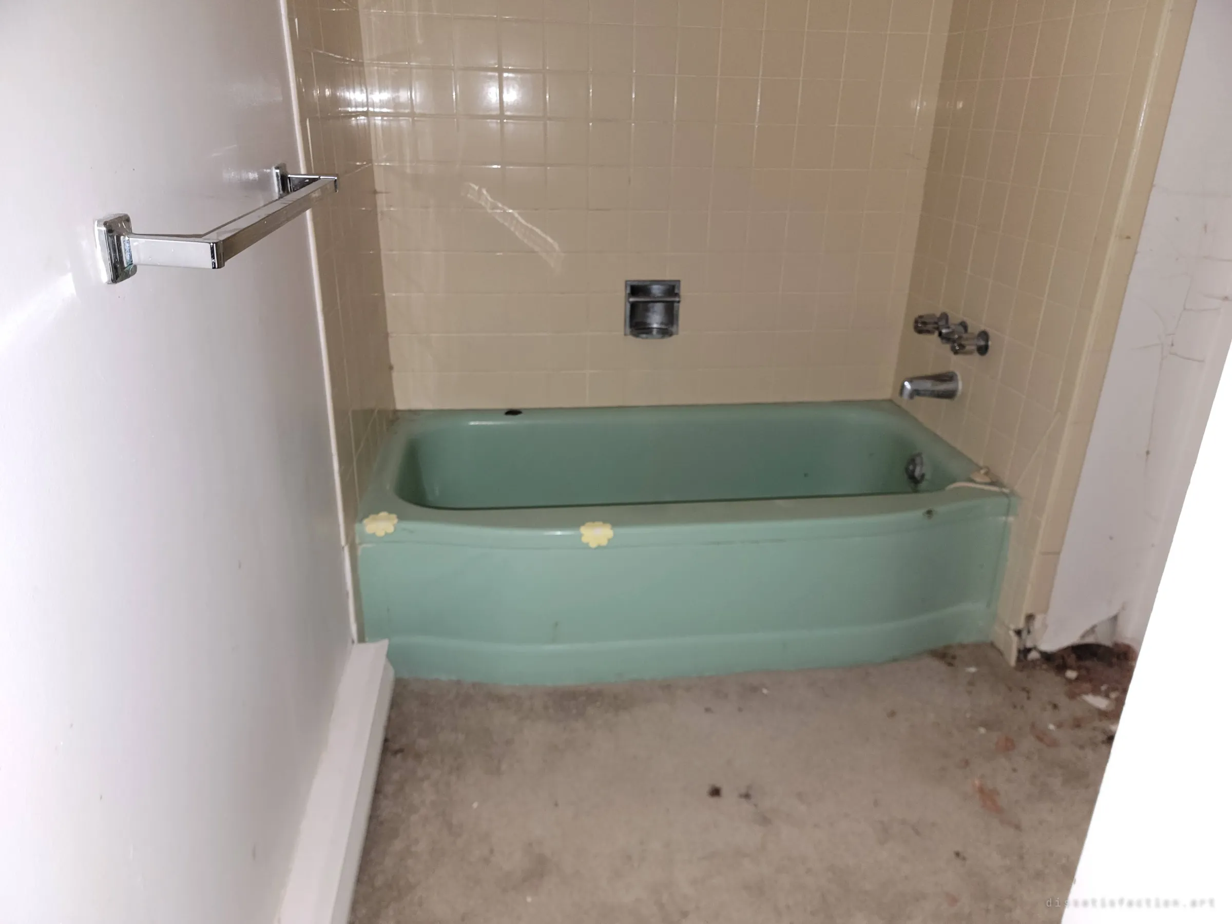 A sea-green bathtub with daisy anti-slip stickers set in beige tile. A preserved mid-century detail amid the ruin.