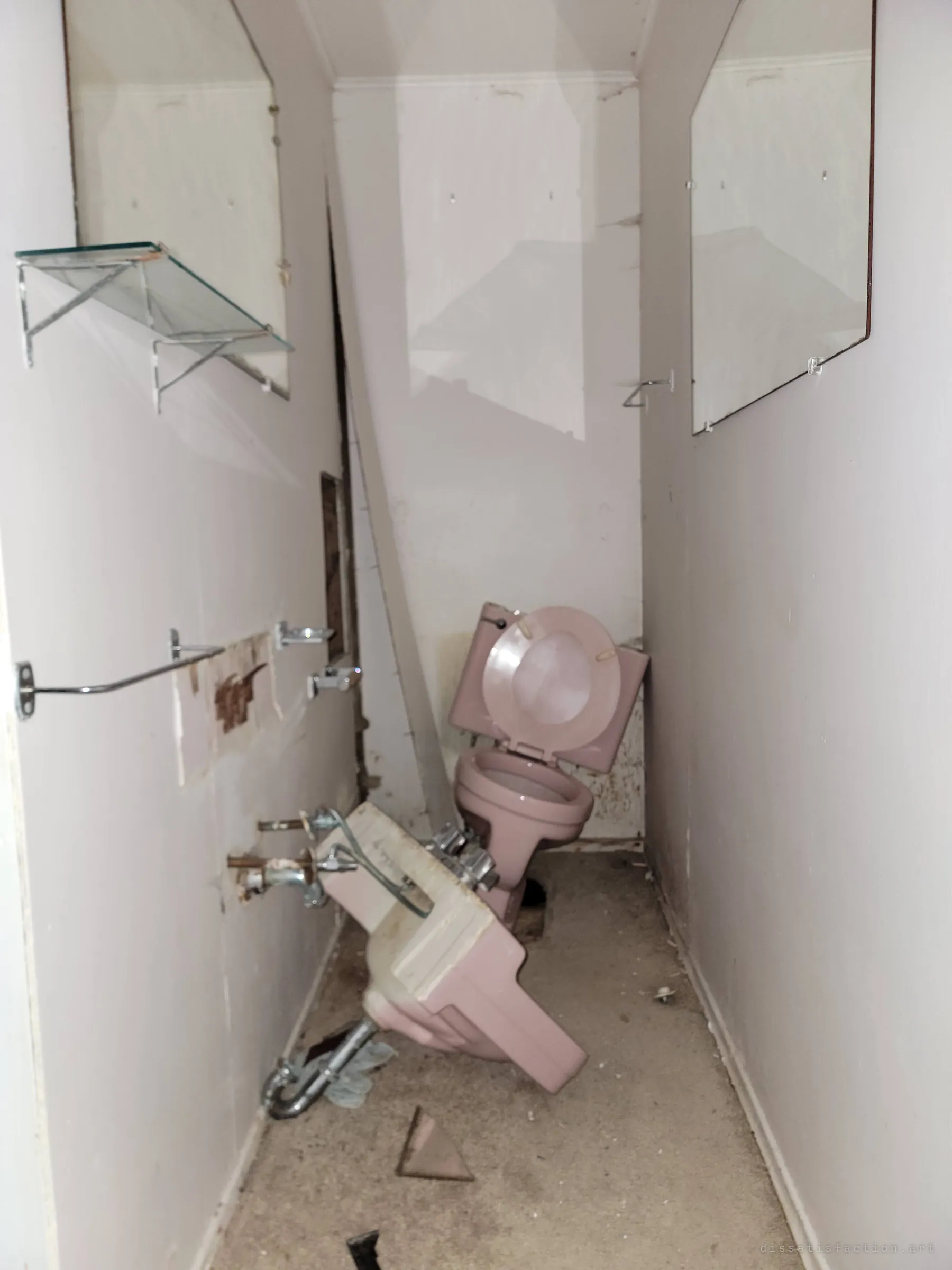 A narrow bathroom where a pink sink has been ripped from the wall and the toilet knocked askew between two mirrors. The damage shows the vandalism the house suffered.