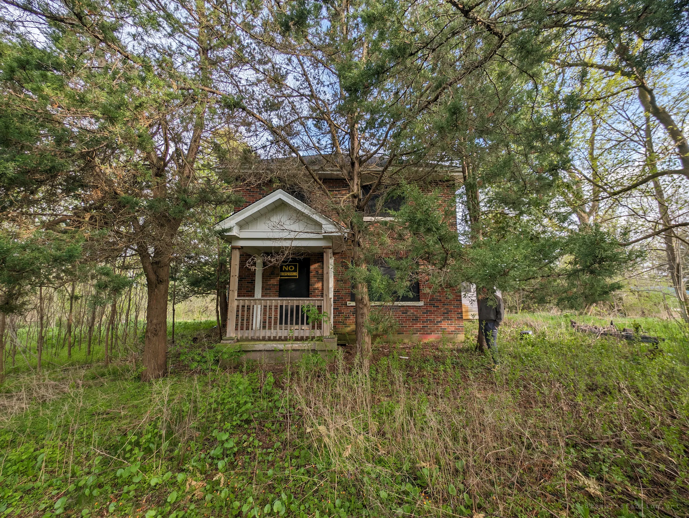 Overgrown red-brick farmhouse half hidden behind cedars, a no-trespassing sign on the boarded front door and a companion standing by a tree. It sets the scene: the house is being swallowed by the yard.