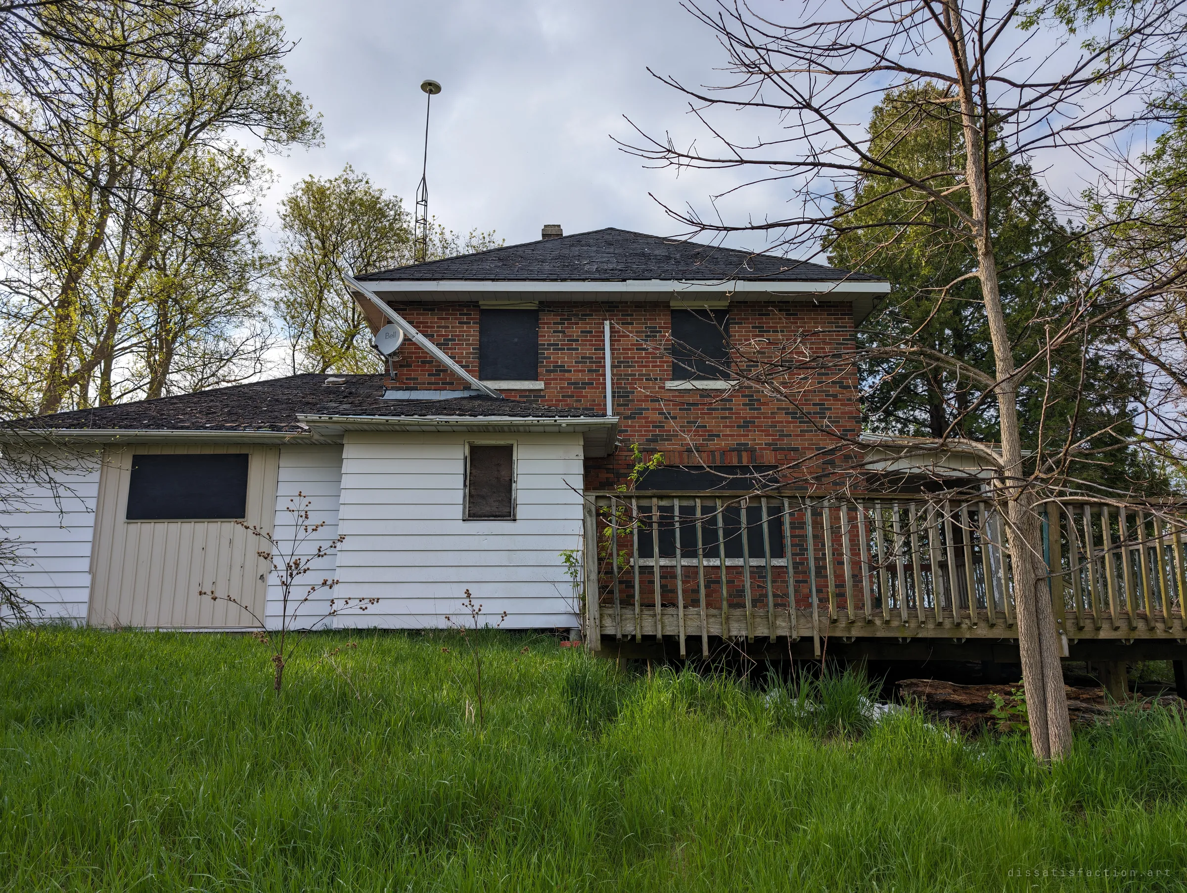 Rear of the brick house with a white-sided addition, boarded windows, a sagging deck and a tall antenna mast above the roof. The two-tone brick pattern gives the album its name.