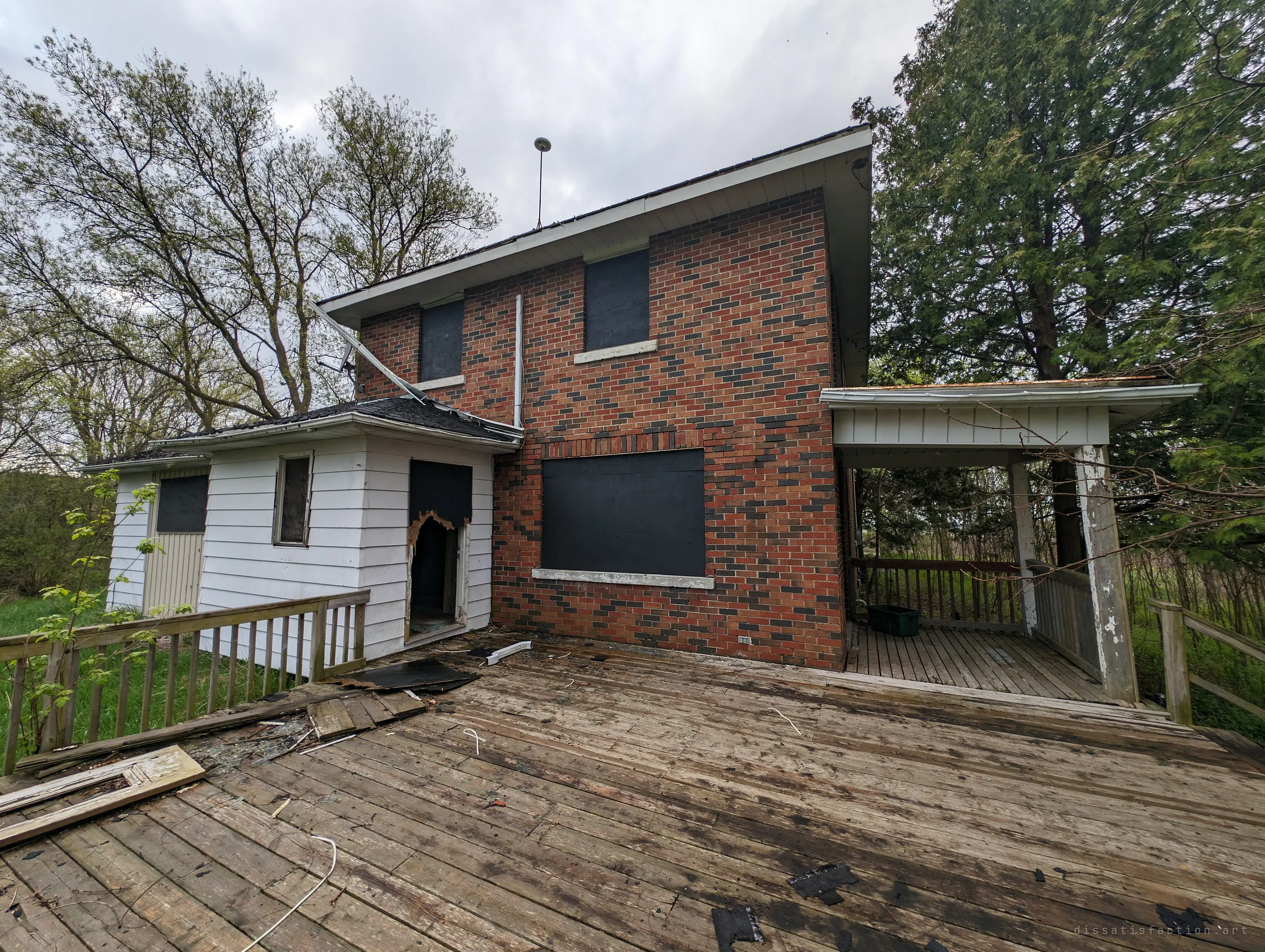 Back deck strewn with shingles and debris, the addition door burned open and every window boarded. Scorch marks hint at a past fire.