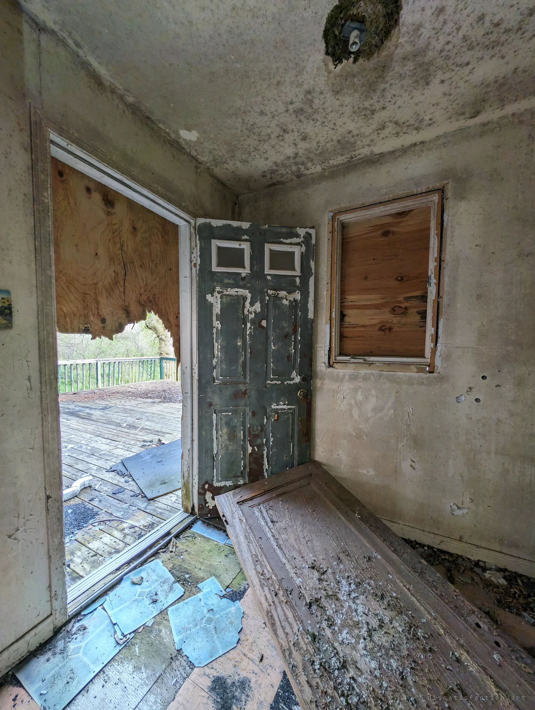 Mould-speckled entry room with a peeling green door hanging open onto the deck and a loose door lying across broken floor tiles.