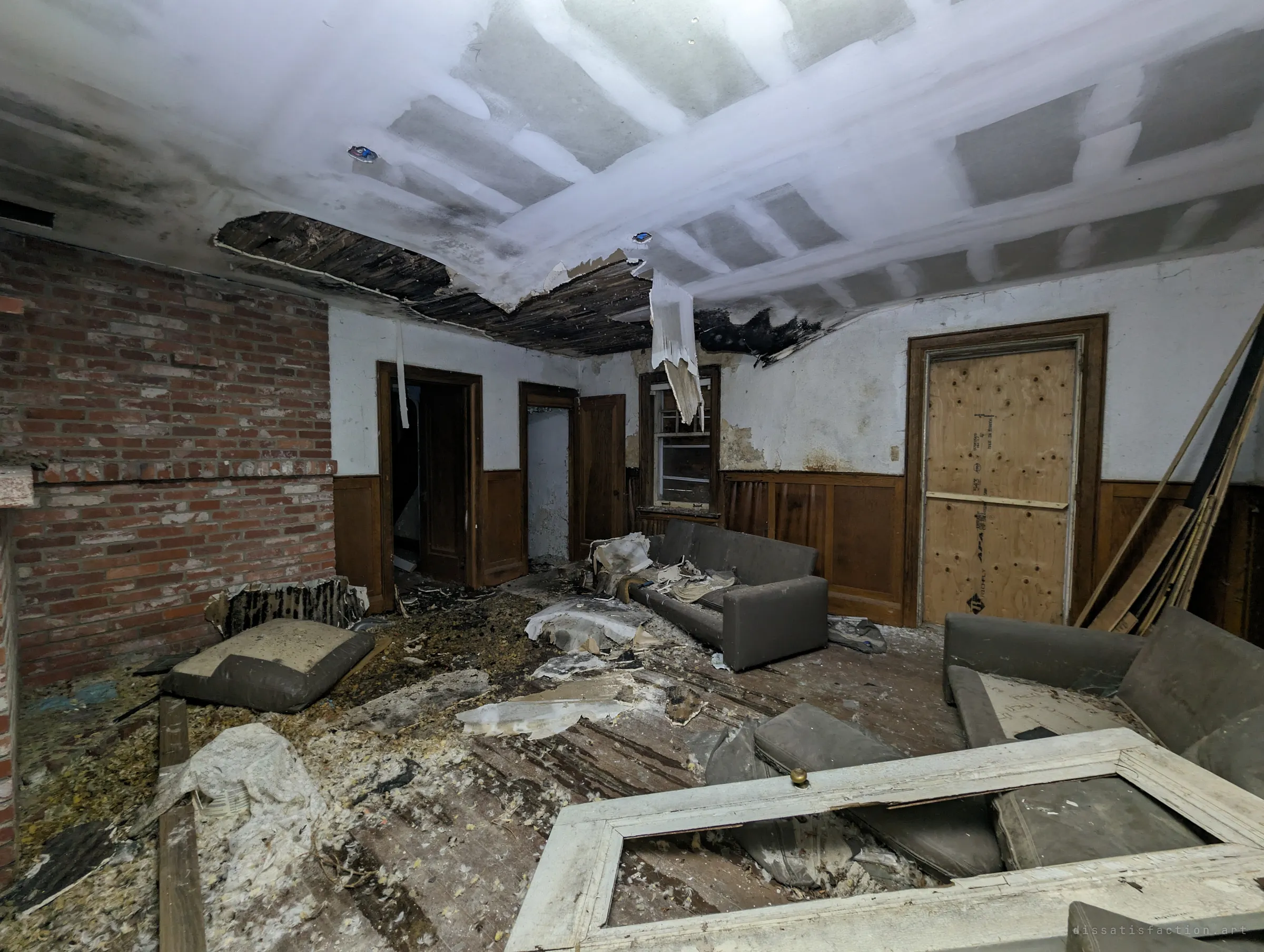 Living room under a collapsing, water-stained ceiling, with a brick fireplace wall, grey couches and debris scattered across the wood floor.