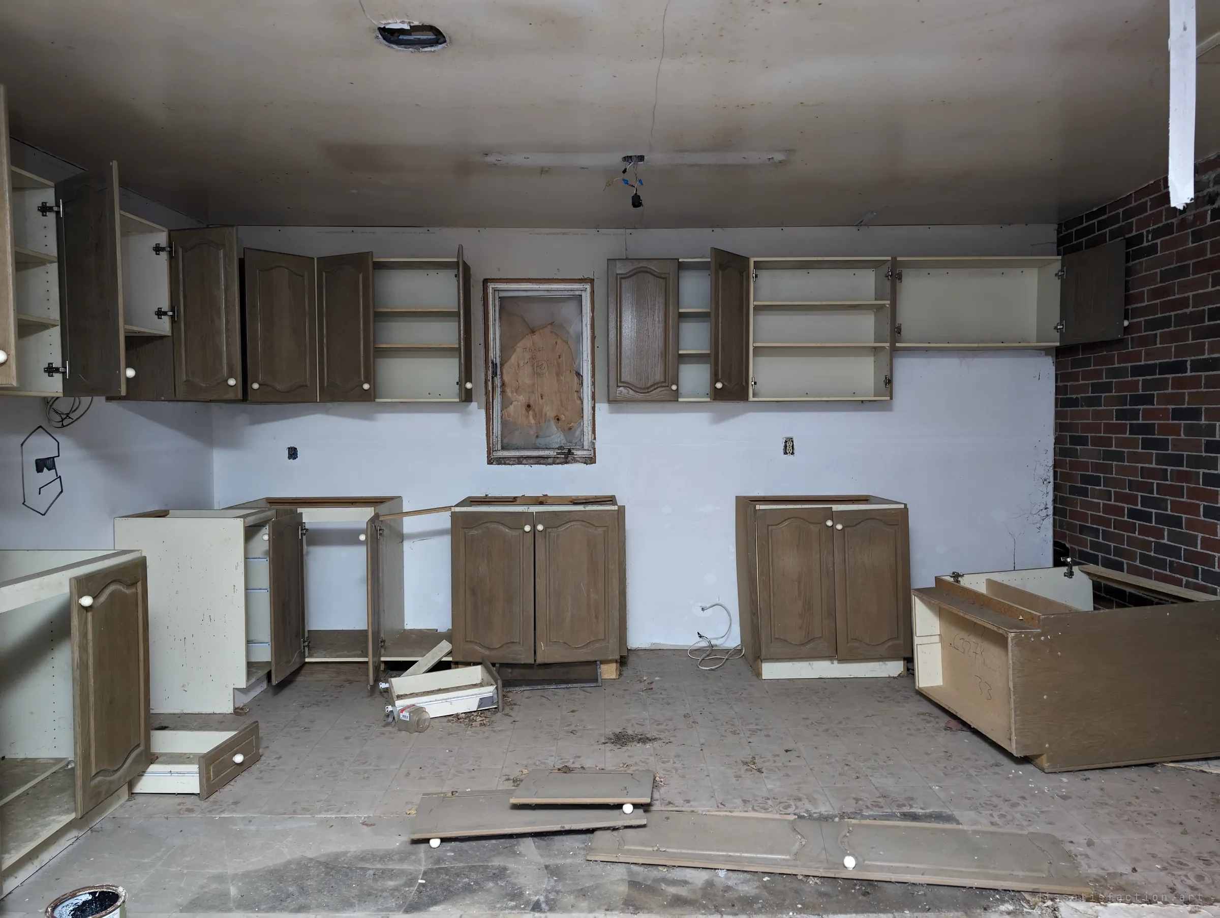Stripped kitchen with cabinet doors hanging open and base units pulled from the wall, a boarded window set between the upper cabinets.