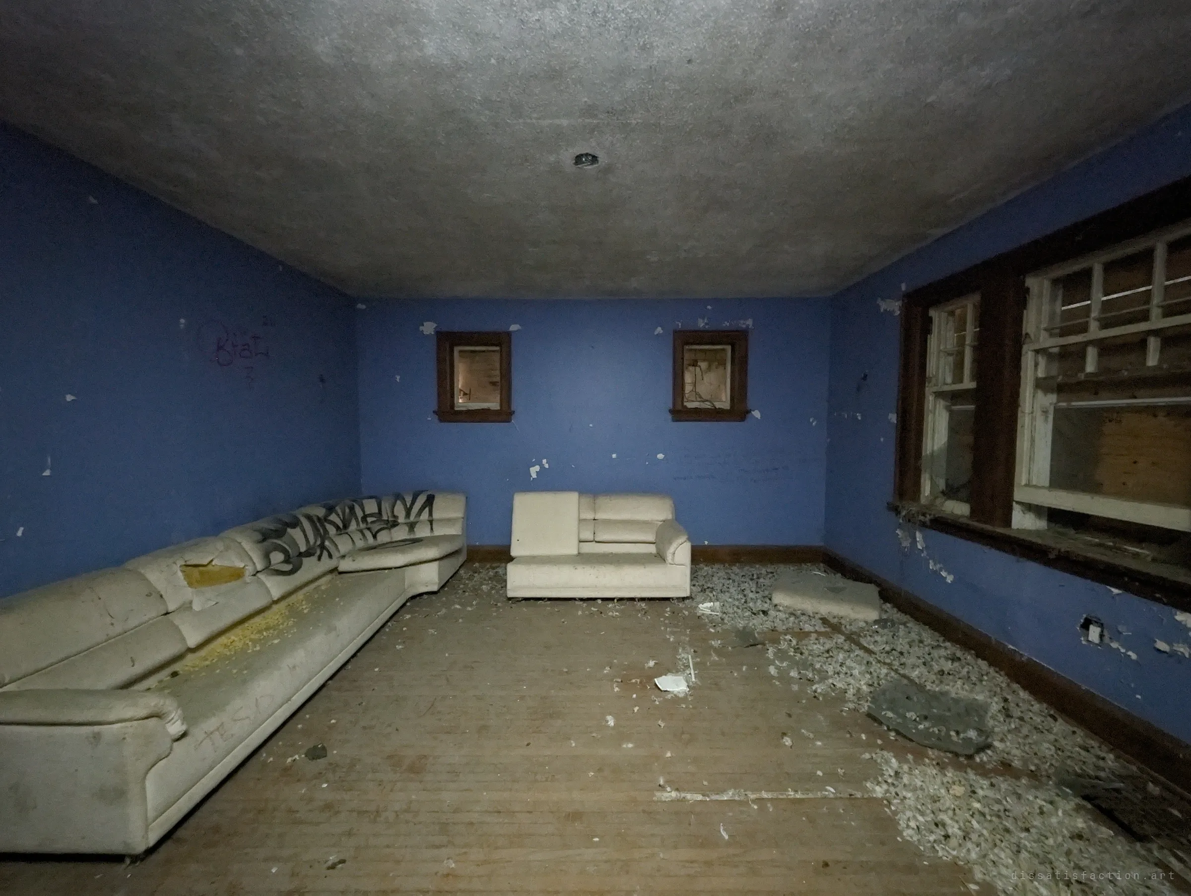Periwinkle-blue lounge with a graffiti-tagged cream sectional and insulation spread across the floor below boarded windows.