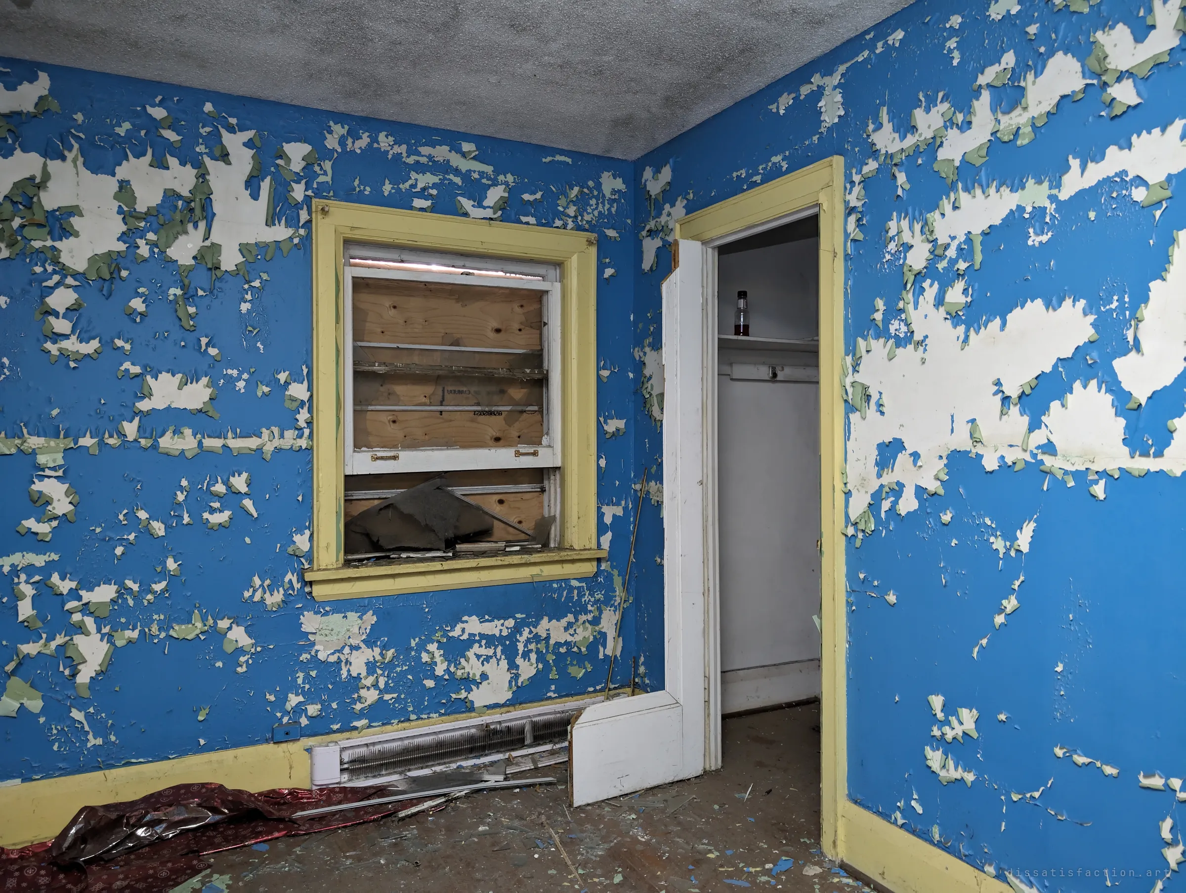 Small room with bright blue paint peeling off in sheets, a yellow-trimmed boarded window and an open closet with a lone bottle on the shelf.