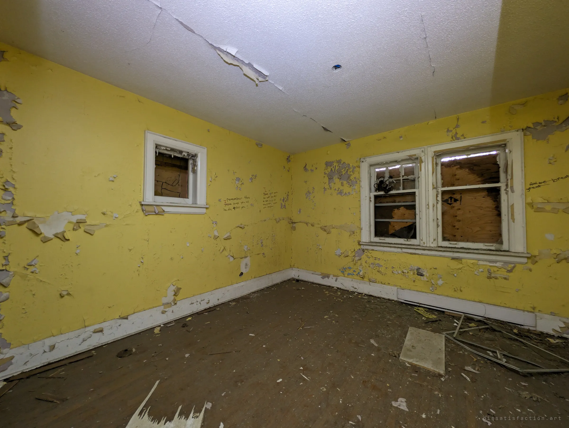 Yellow bedroom with flaking paint, cracked ceiling and handwritten messages on the wall between two boarded windows.