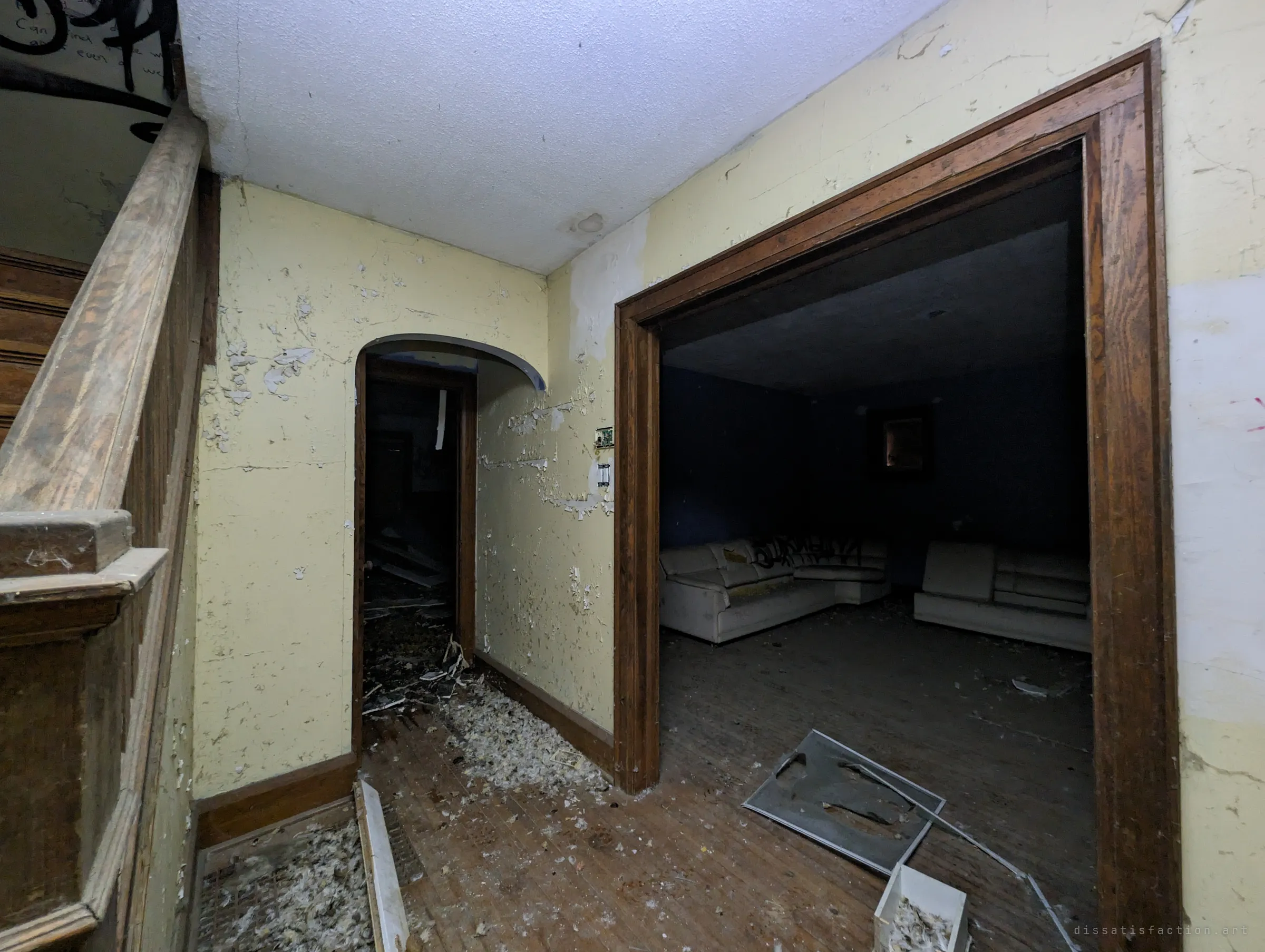 Front hall with an oak banister and an arched doorway, looking through a wide wood-framed opening into the dark blue lounge with its tagged sectional.
