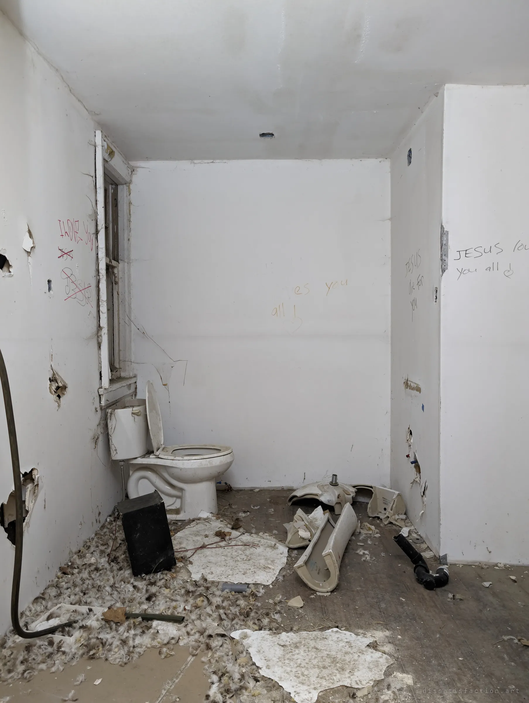 White bathroom with a lone toilet, a smashed sink on the floor, feathers everywhere and handwritten religious messages on the walls.