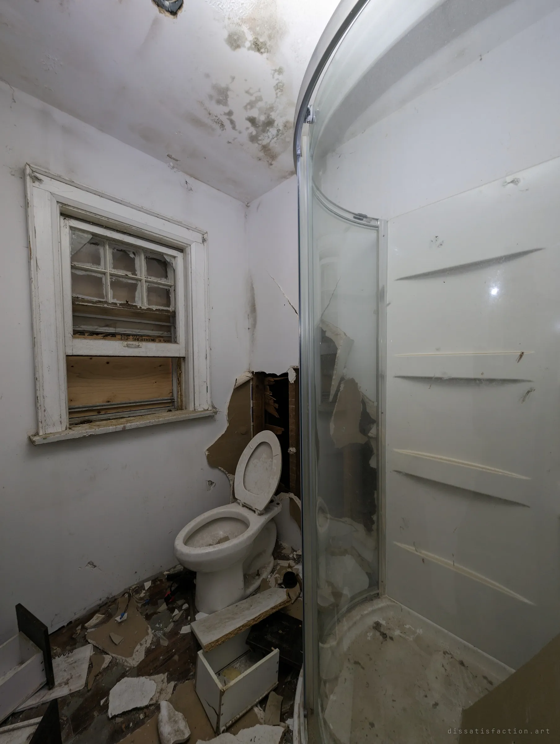 Cramped bathroom with a mould-stained ceiling, a torn-open wall behind the toilet and a curved glass shower stall.