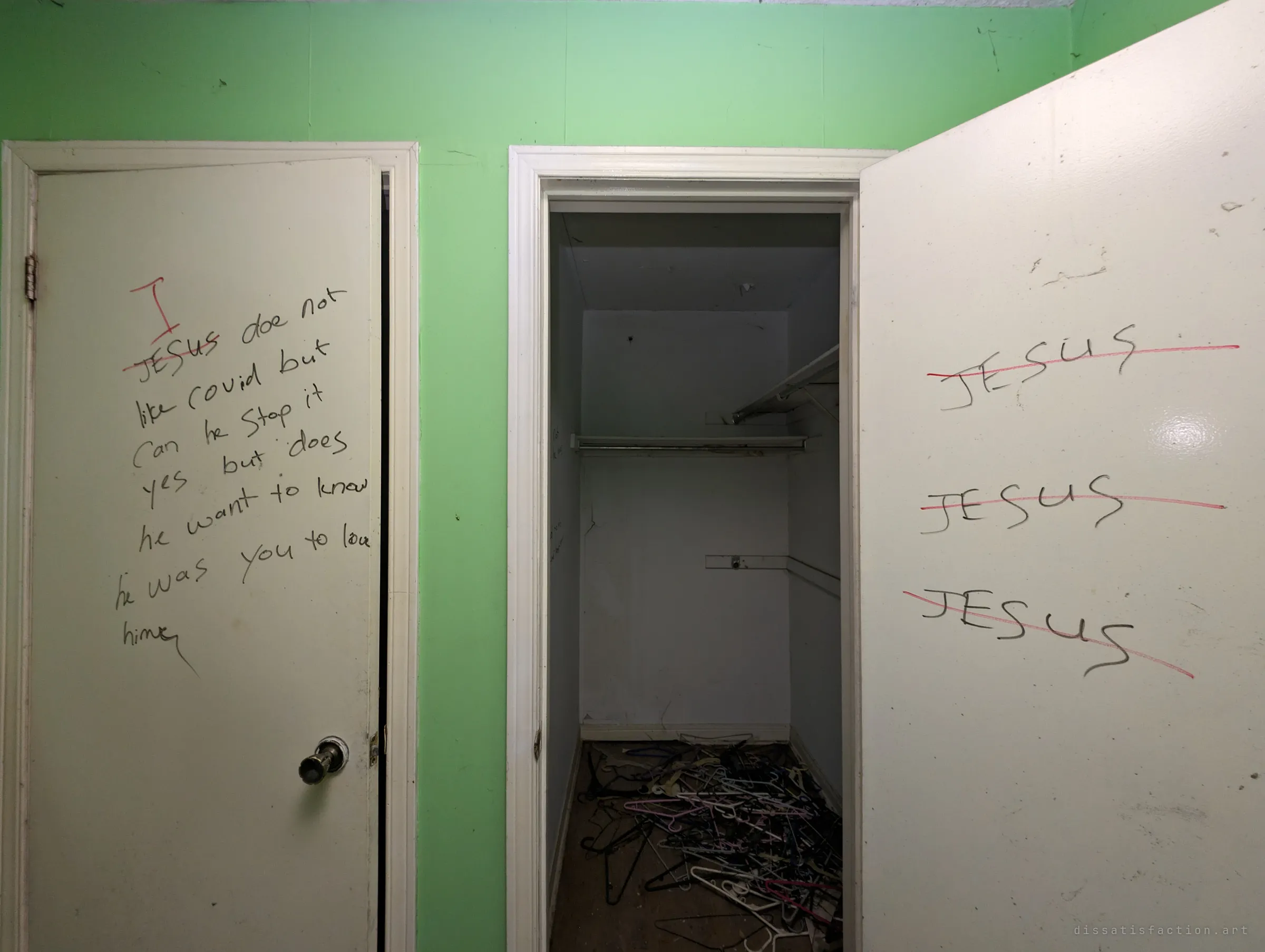 Two white doors on a mint-green wall covered in marker preaching, every written name crossed out in red and replaced with 'I'; between them an empty closet holds a heap of tangled coat hangers. The obsessive rewriting sets the unsettling tone of the house.