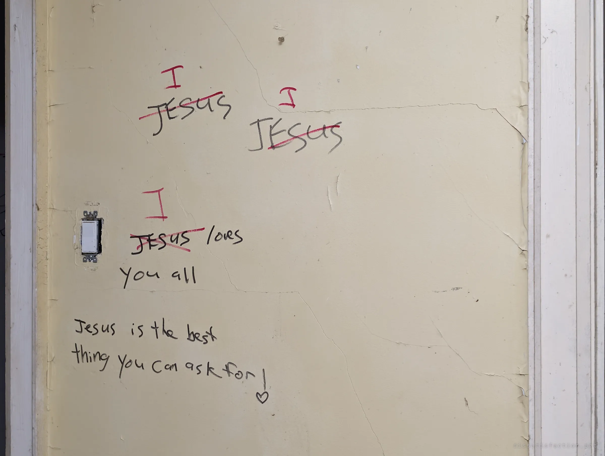 A cracked cream wall beside a light switch carries handwritten devotional lines, with the name crossed out in red and an 'I' written above each one. A small heart closes the message.