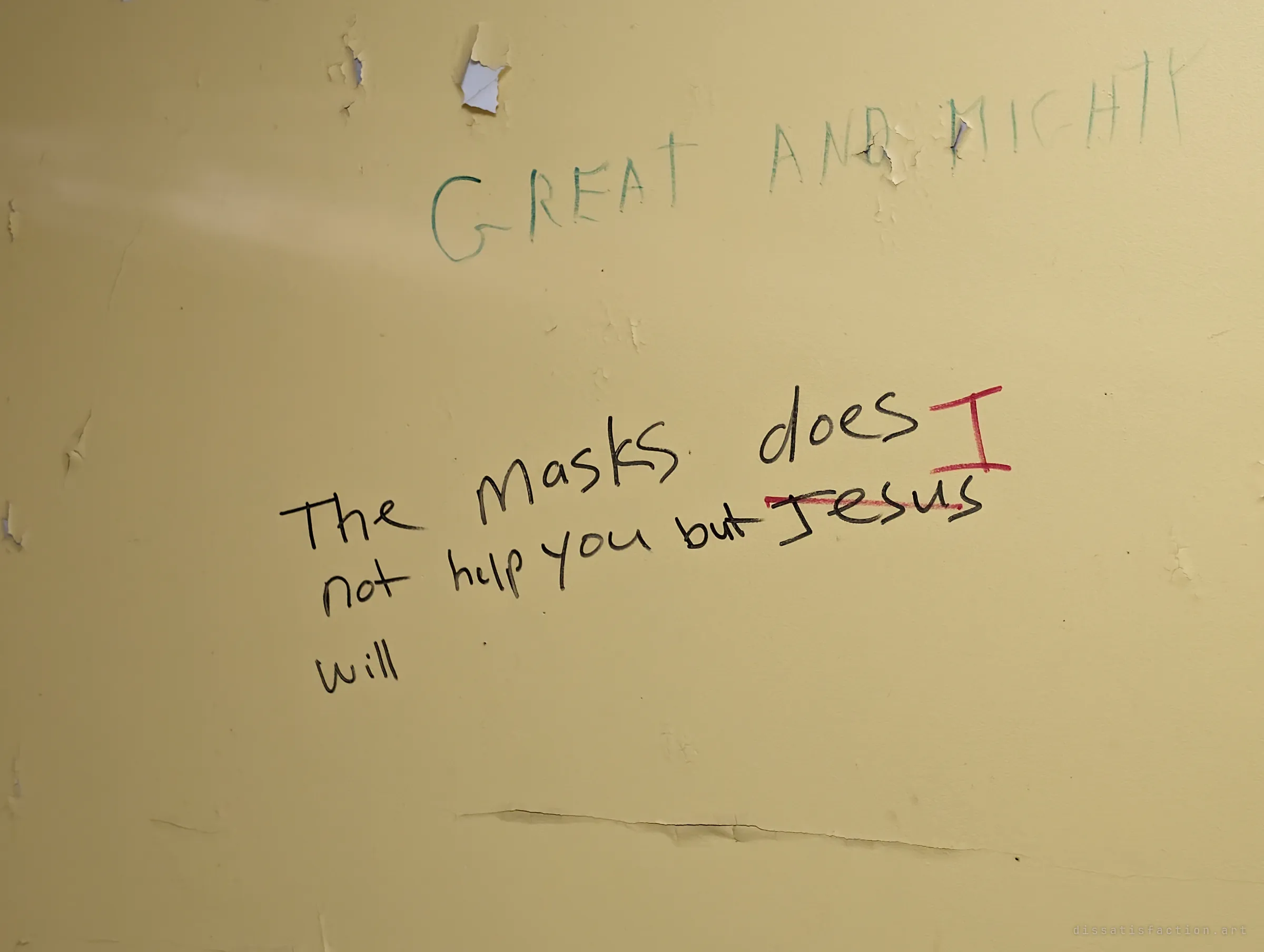 Peeling yellow wall with faded green capital letters praising the Lord and a black marker note saying masks will not help, its name again corrected in red to 'I'. A pandemic-era message left behind in an empty room.