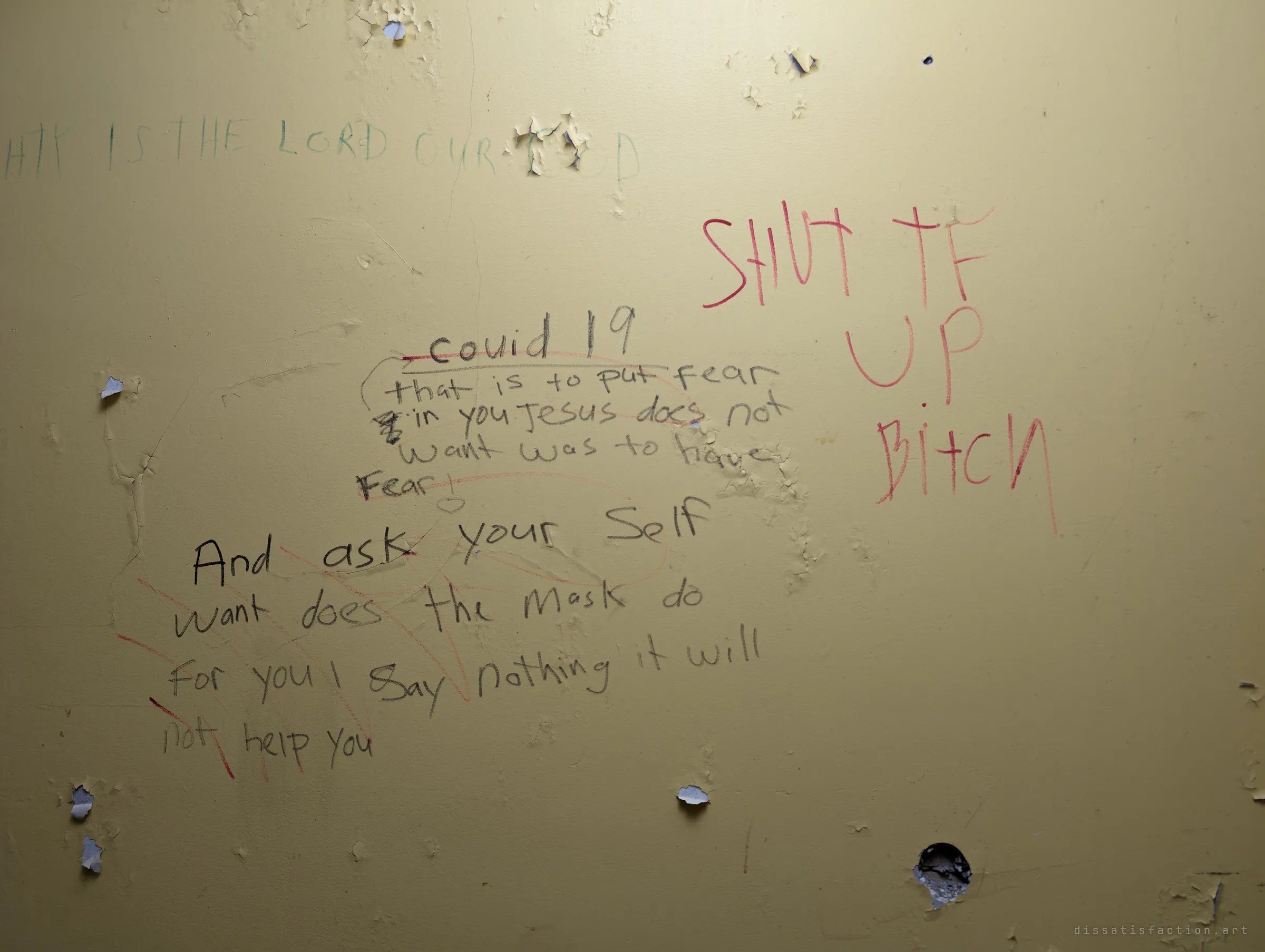A yellow wall where a long handwritten sermon about covid 19 and masks is answered by a later visitor's large red retort, with peeling paint and a punched hole below. Two voices argue across the same surface.