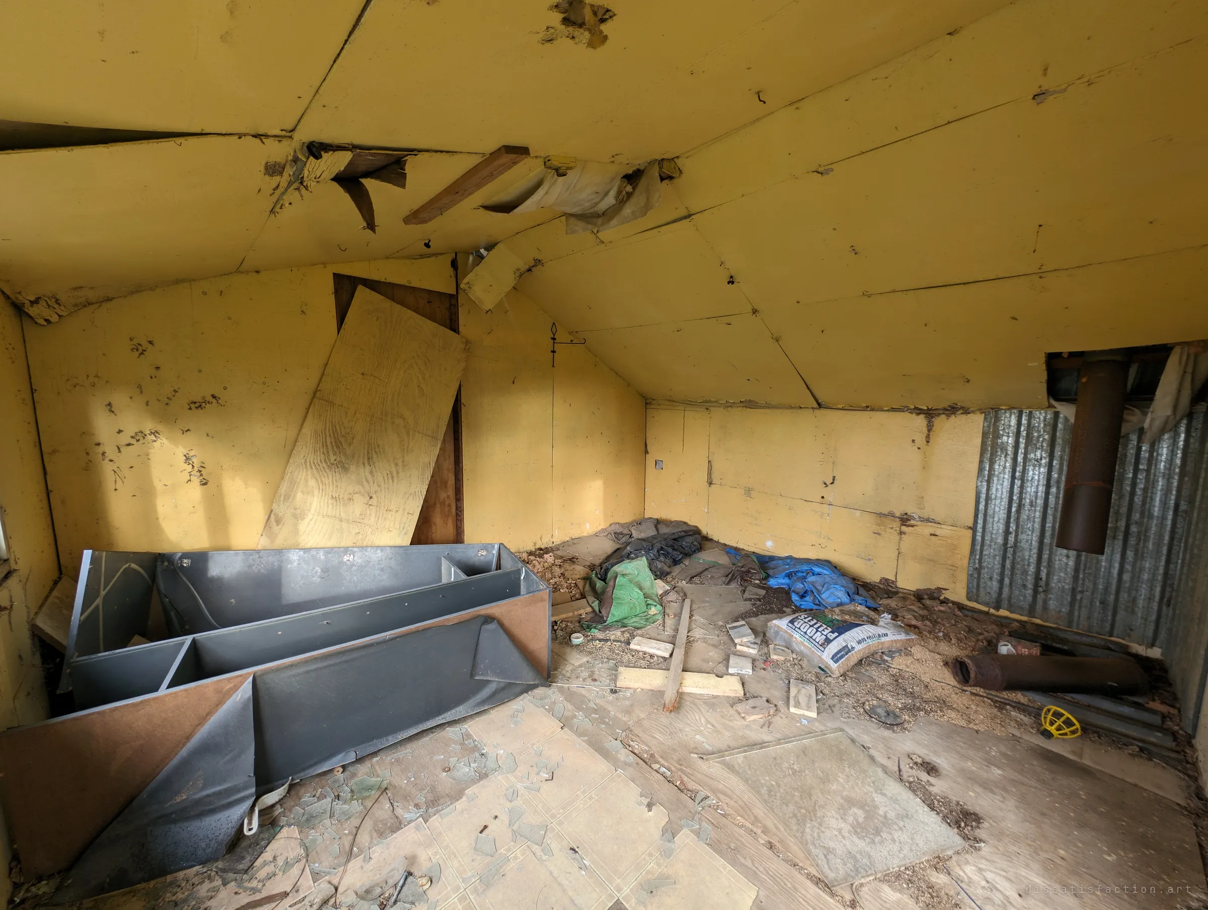 Inside the shed: sagging yellow ceiling panels, a toppled grey shelving unit, broken glass, tarps and a sack of wood pellets beside a rusted stovepipe. The small room has been left to rot from the roof down.